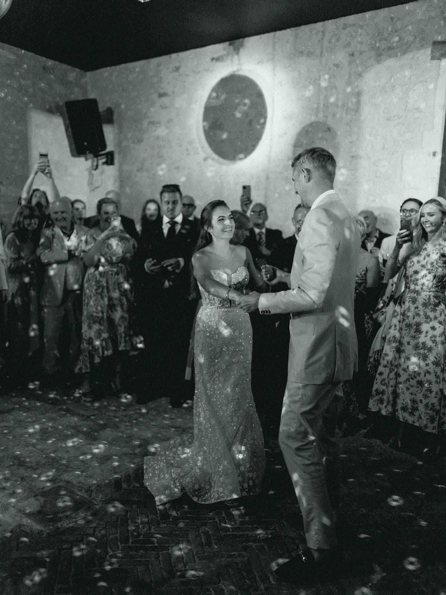 This black-and-white image captures a couple's first dance at an indoor reception venue with exposed stone walls and a herringbone brick floor. The bride wears a strapless, heavily embellished full-length gown with visible sequin or beaded detailing, while the groom is dressed in a light-colored suit with no tie and his shirt collar open. Soap bubbles fill the air and are scattered across the floor, adding a festive element to the moment. Approximately 20 or more guests line the perimeter of the dance floor, many holding up phones to photograph or film, with their expressions showing clear enjoyment; the image has strong contrast with deep shadows in the foreground and a warm spotlight illuminating the stone wall behind the couple.