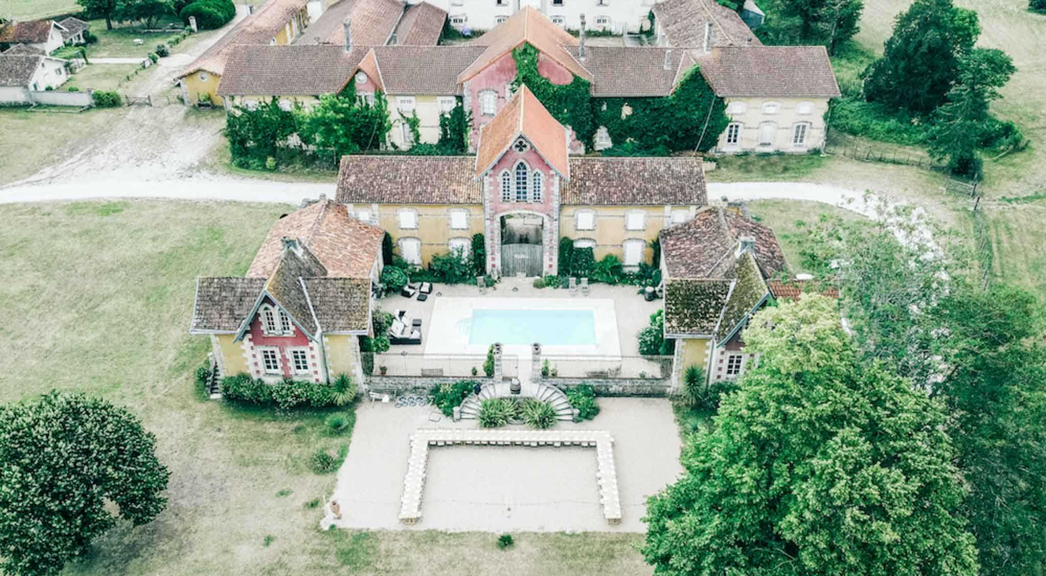 An aerial drone shot of a French country estate venue, showing the full property layout from above. The complex features a central building with a distinctive Gothic-arched gateway entrance in red brick, flanked by two symmetrical pavilions with terracotta-tiled roofs, and a rectangular swimming pool in the central courtyard. In the foreground, a ceremony space is visible with rows of white chairs arranged in a U-shape on a gravel terrace, connected to a formal garden area with a circular ornamental feature. The surrounding grounds include open fields, mature trees, and additional outbuildings with ochre-yellow rendered facades visible in the background. Potential venue feature image.