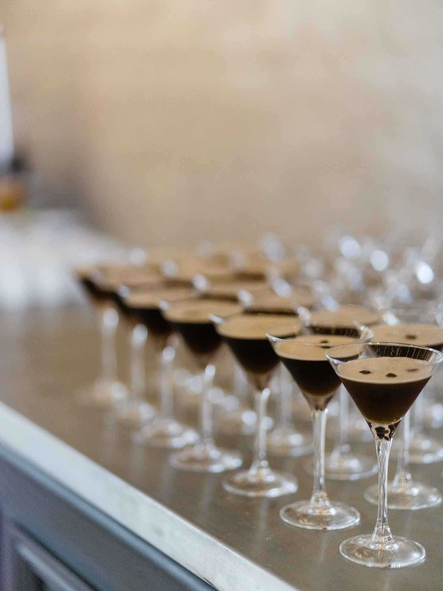 A detail close-up shot of a large batch of espresso martinis lined up on a stainless steel bar counter, ready to be served at a wedding reception. Each martini glass contains a dark brown espresso-based cocktail topped with a light foam layer and three coffee beans as garnish. The image uses shallow depth of field, with the foreground glasses in sharp focus and the rows of glasses behind gradually blurring. A small number of empty glasses are also visible in the background, suggesting ongoing preparation.