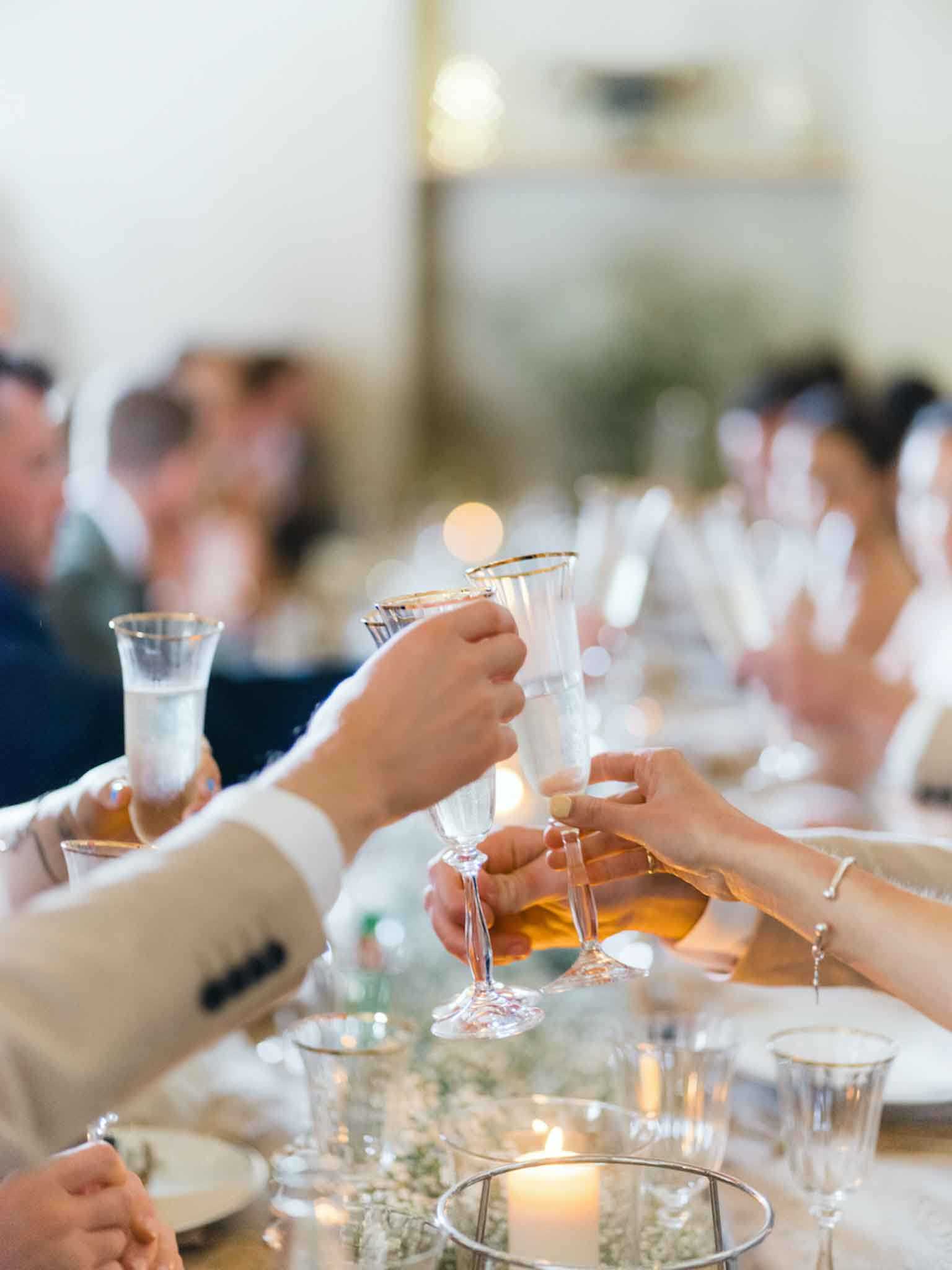 Guests clink crystal champagne flutes with gold rims during a toast at an indoor wedding reception, with multiple hands reaching in from around the table. The setting appears to be a bright, light-filled reception room with blurred guests visible in the background, including someone in a cream suit jacket on the left. The table is decorated with lit pillar candles in glass holders, small white baby's breath floral arrangements, gold-rimmed glassware, and white tableware, suggesting a classic and refined decor palette. The shot is a close-up, shallow depth-of-field composition focused on the clinking glasses with the reception scene softly blurred behind.