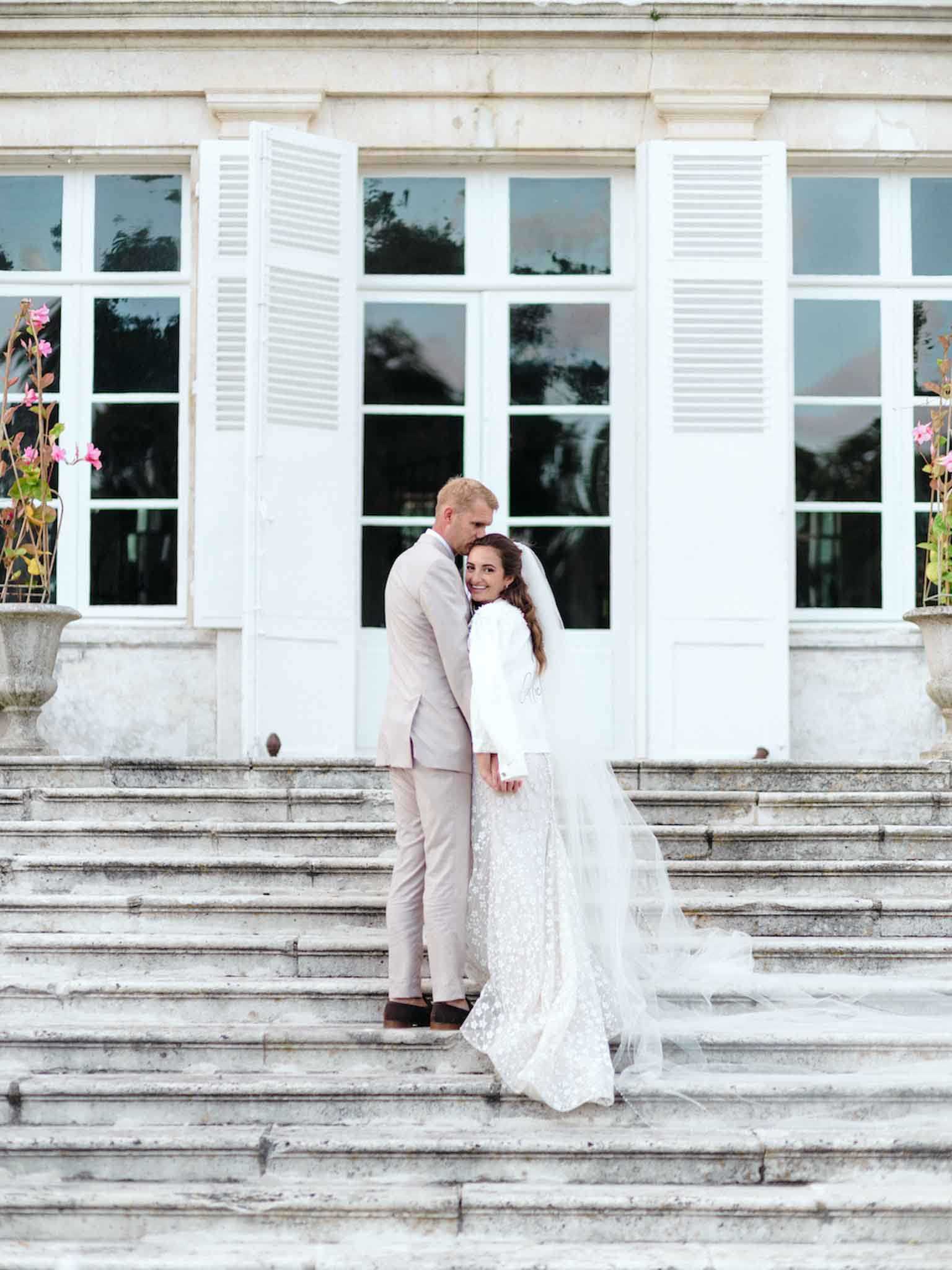 A couple portrait taken on the wide stone staircase of a French chateau or manor house, with tall white-shuttered French doors and stone balustrade planters with pink climbing flowers visible in the background. The bride wears a long-sleeved ivory lace gown with floral embroidery and a cathedral-length veil that trails down the steps, while the groom wears a light sand/greige slim-fit suit with dark brown shoes. The groom leans in to kiss the bride's cheek as she turns toward the camera smiling, creating a candid couple moment. This is a full-length portrait shot with the architectural facade centered behind the couple. Potential venue feature image.