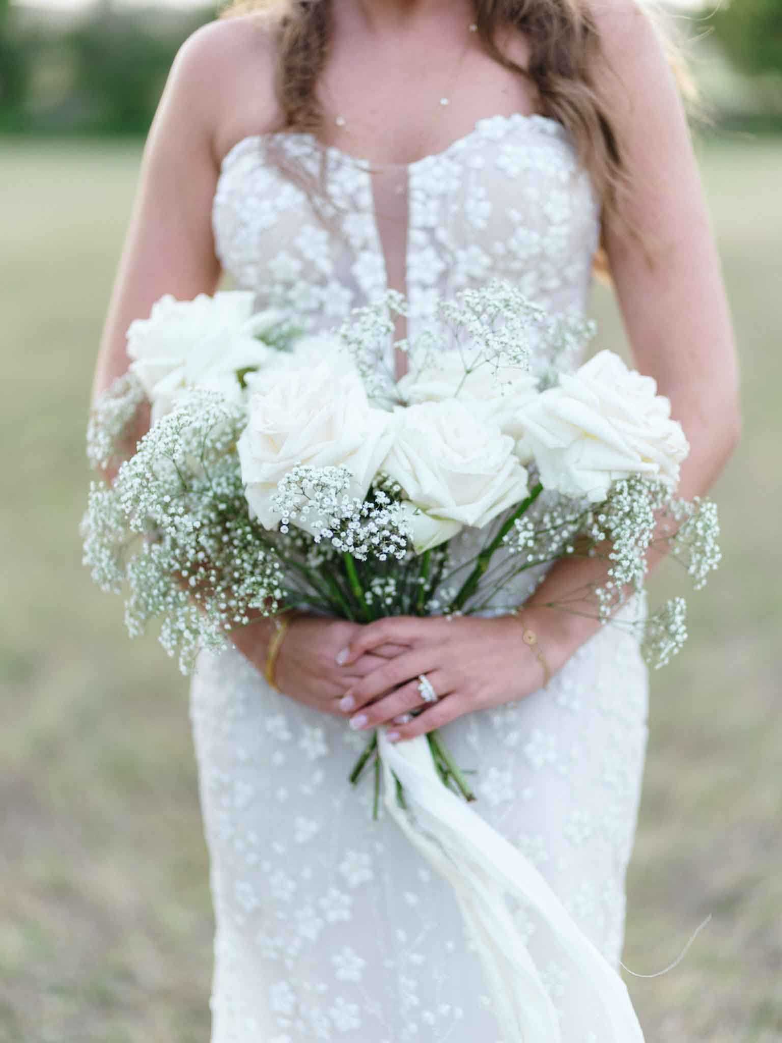 A close-up portrait of a bride standing outdoors in an open field, cropped from the neck to below the hips. She wears a fitted white lace gown with a floral embroidered pattern, a sheer plunging neckline panel, and a sweetheart silhouette. She holds a bridal bouquet composed of white roses and abundant baby's breath, tied with a white ribbon streamer. A diamond engagement ring and a delicate gold bracelet are visible on her hands. Her long wavy hair falls over her shoulders. The background is softly blurred with a neutral, muted green tone, keeping full focus on the dress and bouquet.