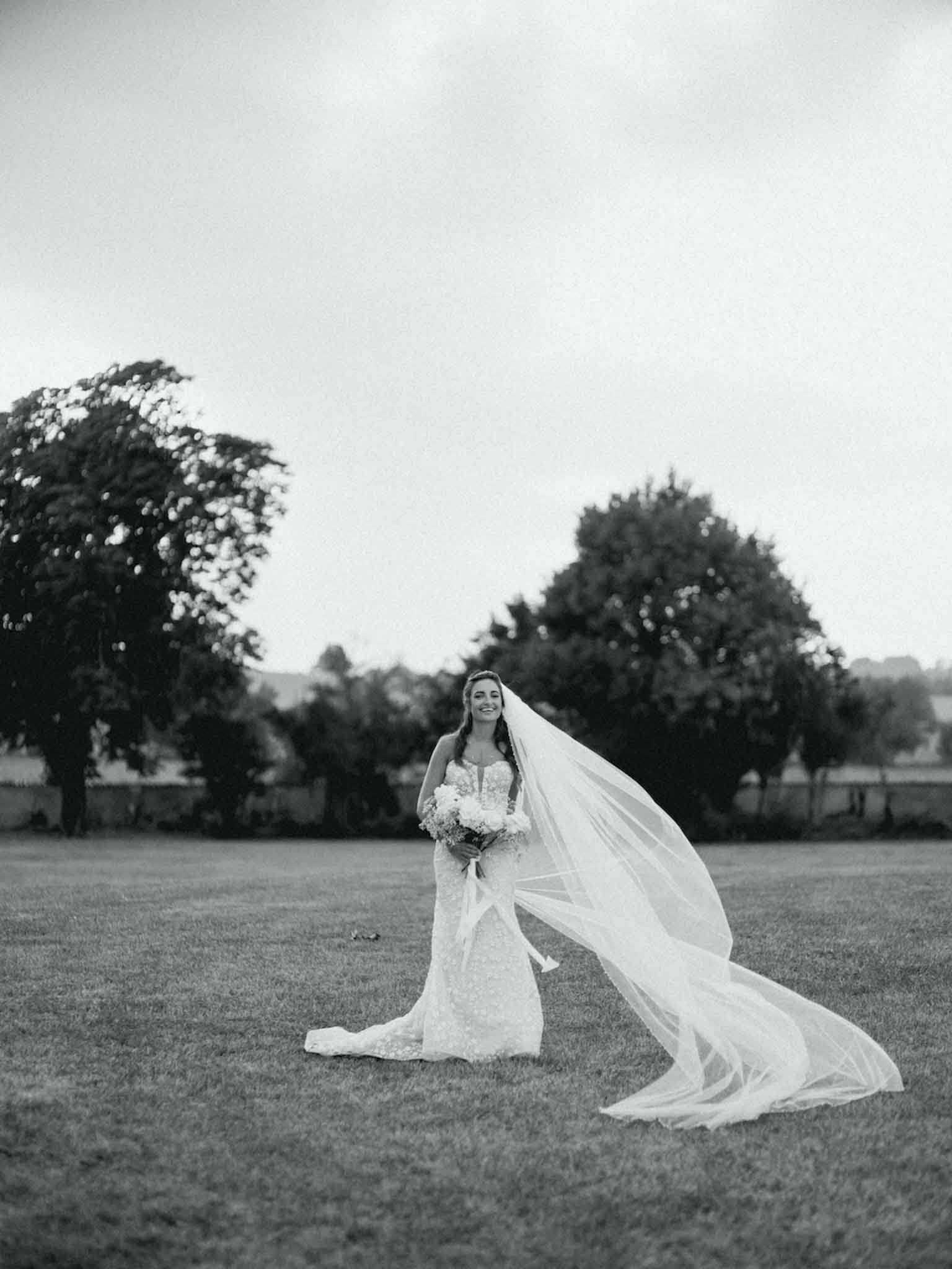 This is a black-and-white bridal portrait taken outdoors on an open lawn, with trees and what appears to be a low building visible in the background. The bride stands alone in the center of the frame wearing a fitted lace gown with a sweetheart neckline and a long cathedral-length veil that is caught in the wind, extending dramatically to the right of the frame. She holds a round bouquet of light-toned flowers and is smiling broadly. The image is a full-length wide shot with high contrast between the bright veil and the mid-tone tones of the lawn and trees.