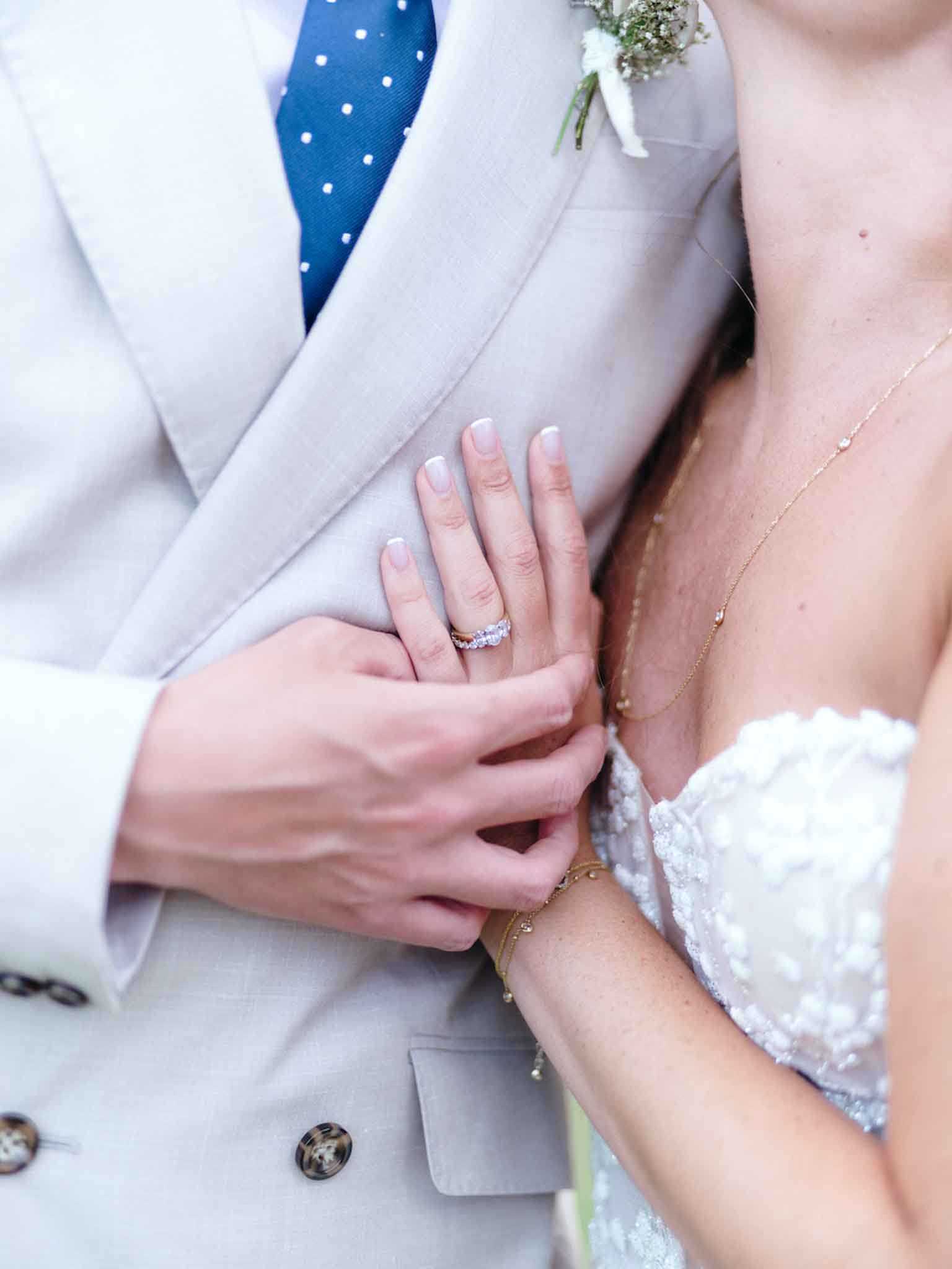 A close-up detail shot of a bride and groom's hands, with the bride's hand resting on the groom's arm as he holds her wrist. The bride wears a diamond eternity band with French-manicured nails, a delicate gold chain bracelet, and a fine gold necklace with small diamond accents visible at her décolletage. She is wearing a strapless white lace wedding dress. The groom is dressed in a light grey double-breasted suit with a navy blue polka-dot tie and a white baby's breath boutonnière. The composition is a tight detail/close-up crop showing only the torsos and hands of the couple.