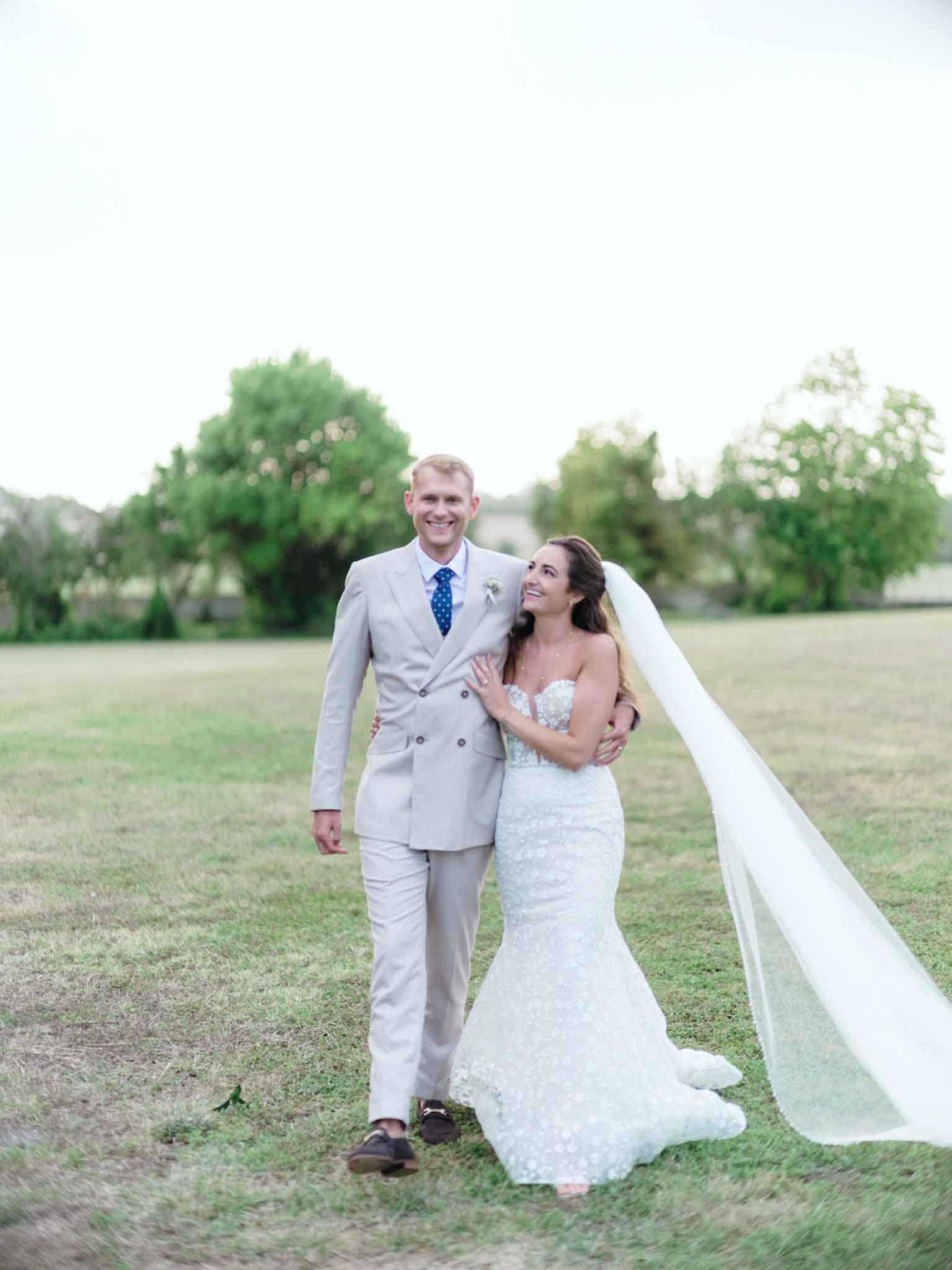A couple portrait taken outdoors on an open lawn, with the bride and groom walking together toward the camera. The groom wears a light beige double-breasted suit with a navy blue polka-dot tie and dark brown loafers, with a small white boutonniere on his lapel. The bride wears a fitted strapless mermaid-silhouette gown in white with an allover floral lace or appliqué texture, paired with a long cathedral-length veil that is caught by the wind and flowing to the right. The bride looks up at the groom while he smiles toward the camera, and she rests one hand on his chest. The composition is a full-length portrait shot with a softly blurred background of trees and what appears to be a building in the far distance.