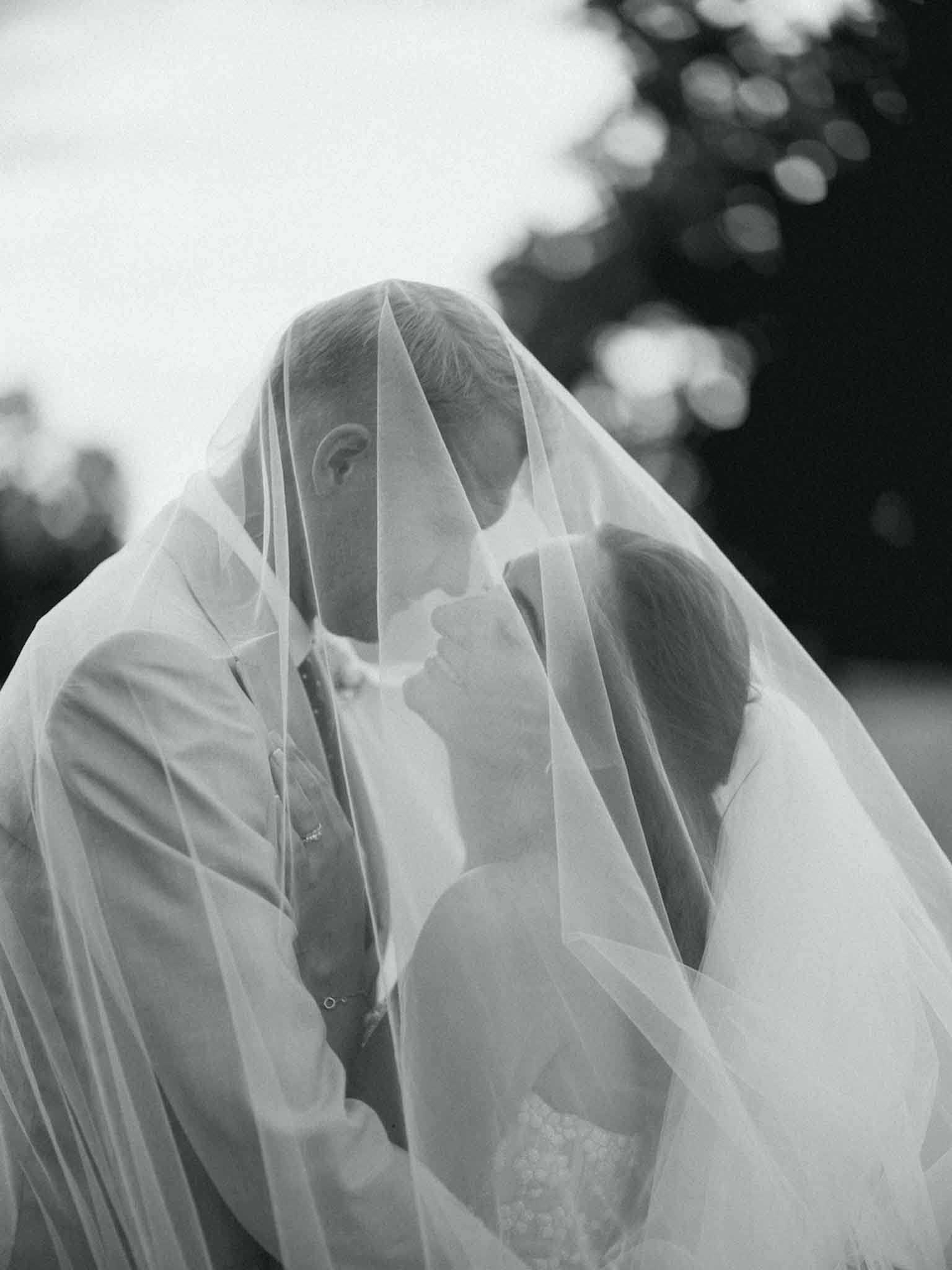 This is a black-and-white portrait of a bride and groom sharing a kiss beneath the bride's long, sheer veil, which drapes over both of them and fills much of the frame. The image is shot outdoors with a softly blurred background and strong contrast between the bright, light tones of the veil and the darker foliage behind. The groom wears a suit with a tie, and the bride's lace dress is partially visible at the lower right. The composition is a close-up portrait emphasizing the layered, translucent texture of the veil as it envelops the couple.