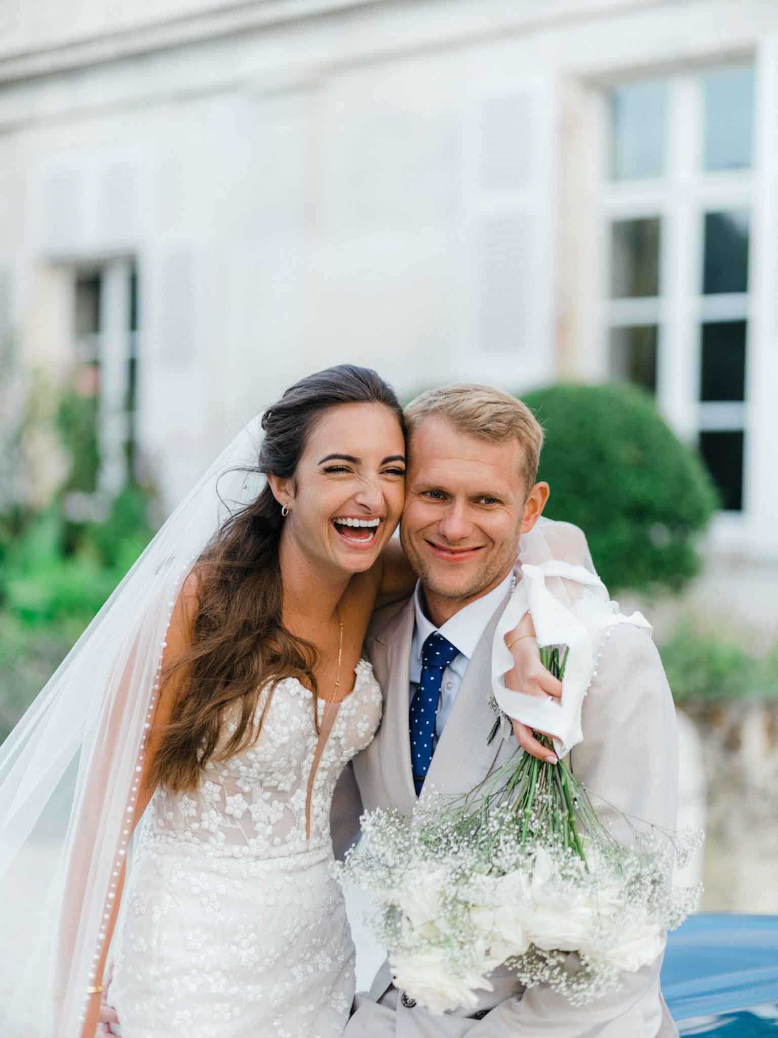 A close-up portrait of a bride and groom posing together outdoors in front of a French chateau facade with white-framed windows visible in the background. The bride wears a fitted ivory lace gown with floral appliqué detailing and a long cathedral veil trimmed with pearl-like edge detailing, and holds a bouquet of white baby's breath and white roses with long green stems wrapped in white ribbon. The groom wears a light grey suit with a white dress shirt and a navy blue polka-dot tie. The bride is laughing openly while leaning her head toward the groom, who smiles directly at the camera. The shot is a mid-length portrait with a shallow depth of field that keeps the couple sharp against the softly blurred chateau exterior.