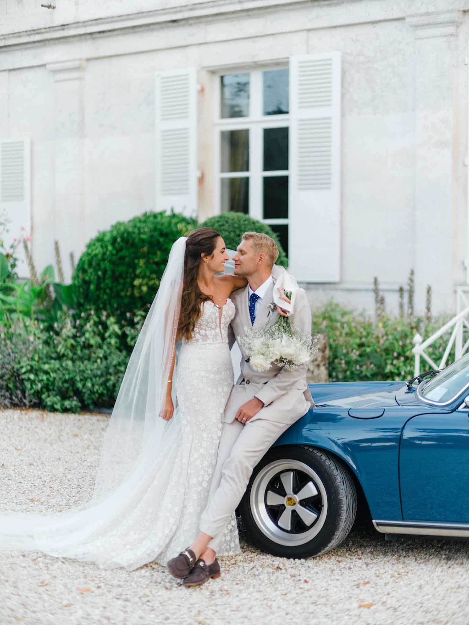 A couple portrait taken outdoors on a gravel driveway in front of a French chateau facade with white shuttered windows. The bride wears a fitted, floor-length lace gown with a sweetheart neckline, a long cathedral-length veil, and brown loafers; she holds a bouquet of white gypsophila and white flowers with trailing greenery. The groom is dressed in a light beige linen suit with a navy polka-dot tie and leans against a vintage blue Porsche 911, holding the bouquet loosely while the couple looks at each other closely. The overall styling is classic-meets-relaxed, with the vintage car serving as a deliberate prop, and the shot is a full-length couple portrait with soft, even natural light.