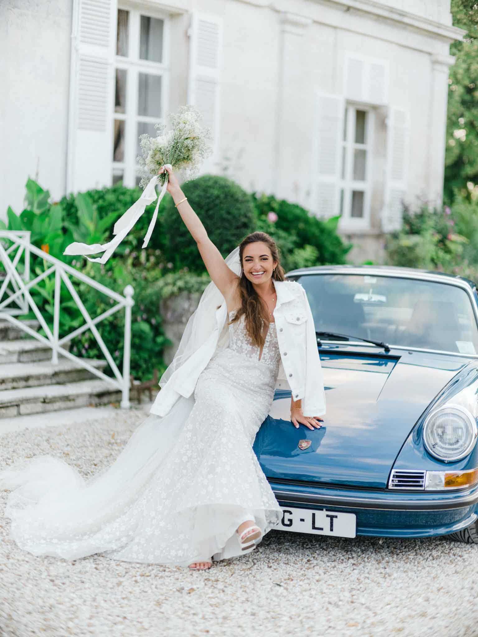 A bride poses leaning against the hood of a vintage blue Porsche 911 on a gravel driveway in front of a French chateau with white shuttered windows. She is wearing a fitted lace wedding gown with a long train, a white denim jacket, and white slide sandals, with a veil worn loosely over her shoulders. Her arm is raised holding a bouquet of white gypsophila tied with long white silk ribbons, and she is smiling broadly at the camera. The composition is a full-length portrait shot with a candid, celebratory feel and a casual-meets-formal styling aesthetic.