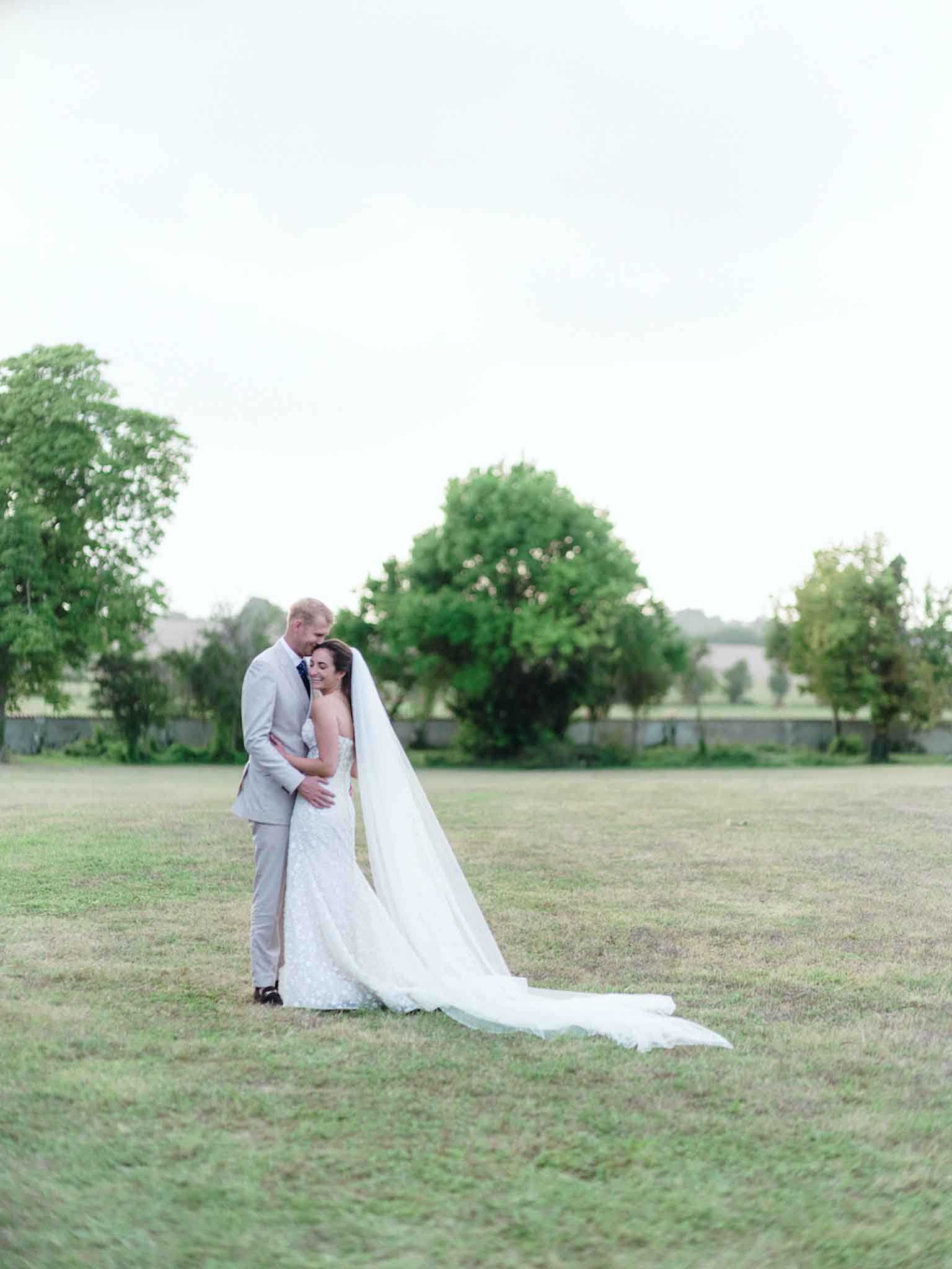 A couple portrait taken outdoors in an open lawn area, with the bride and groom embracing in the center of the frame. The bride wears a fitted, strapless white lace gown with a cathedral-length veil that trails across the ground behind her, while the groom wears a light grey suit with a navy tie. The groom leans his head toward the bride, who smiles with her eyes closed. The composition is a wide shot with significant negative space in the foreground and a low stone boundary wall visible in the background. The overall image has a soft, airy, light-film quality with muted tones.