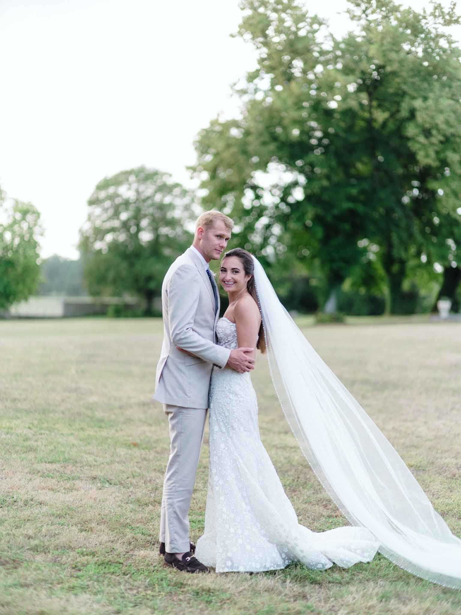 A couple portrait taken outdoors on an open lawn, with the groom standing behind the bride and wrapping his arms around her waist. The groom wears a light beige/sand suit with a dark tie, and the bride wears a fitted strapless gown with allover floral lace or beaded embellishment and a cathedral-length veil that trails dramatically to the right across the grass. The bride looks back toward the camera with a smile while the groom faces forward. The shot is a full-length portrait with a shallow depth of field, set against a backdrop of mature trees in soft evening light.