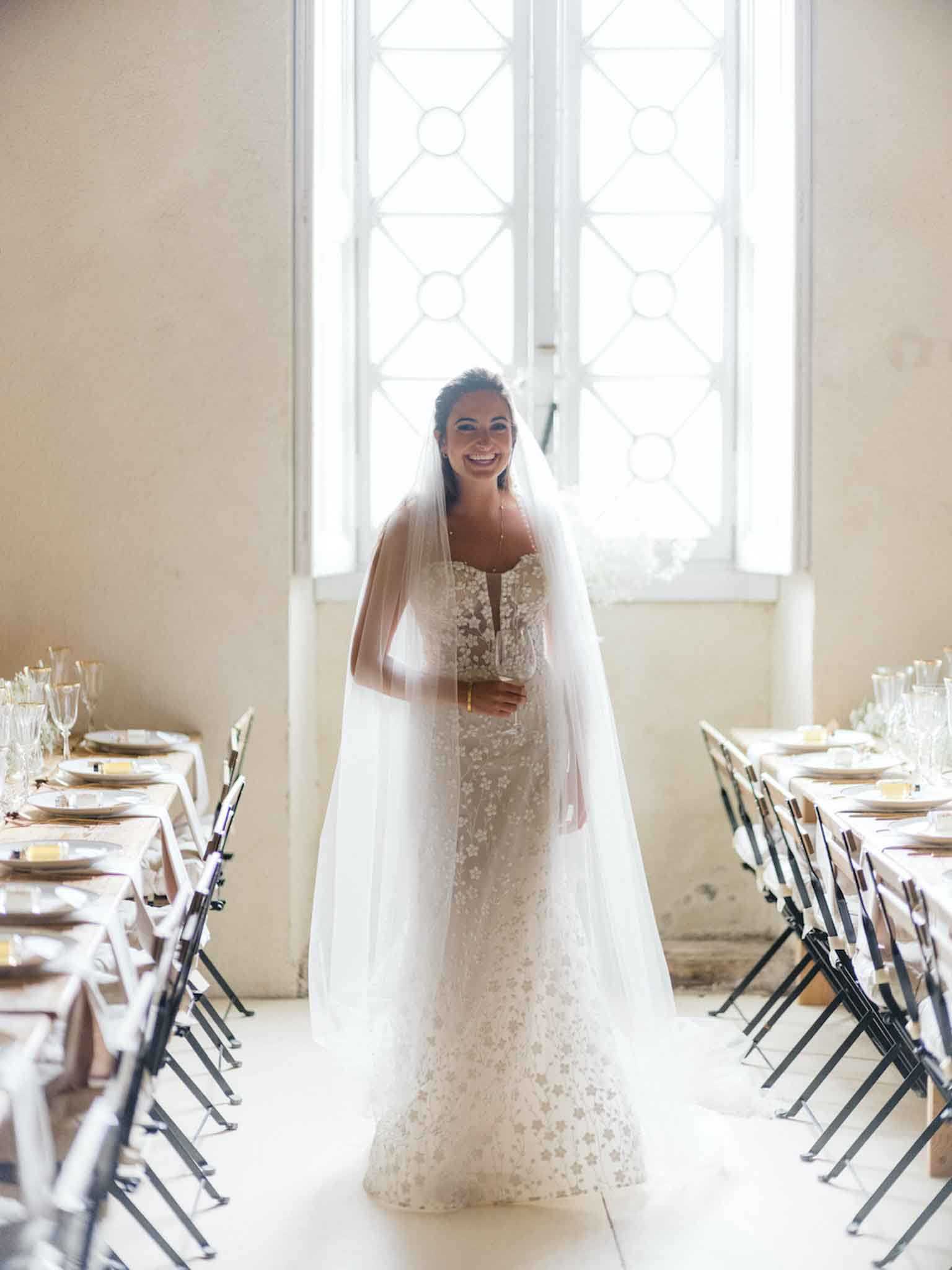 A bride stands in the aisle between two long reception tables inside what appears to be a historic chateau or manor room with tall white latticed windows behind her. She is wearing a fitted ivory lace gown with an all-over floral appliqué pattern and a cathedral-length plain tulle veil, and holds a champagne flute. The reception tables on either side are dressed with white linens, gold charger plates, clear glassware, and black folding chairs with white chair covers. The room has pale, worn plaster walls and light-toned flooring, giving the space a classic French interior feel. This is a full-length portrait shot taken from a slight low angle with natural backlight streaming through the windows.