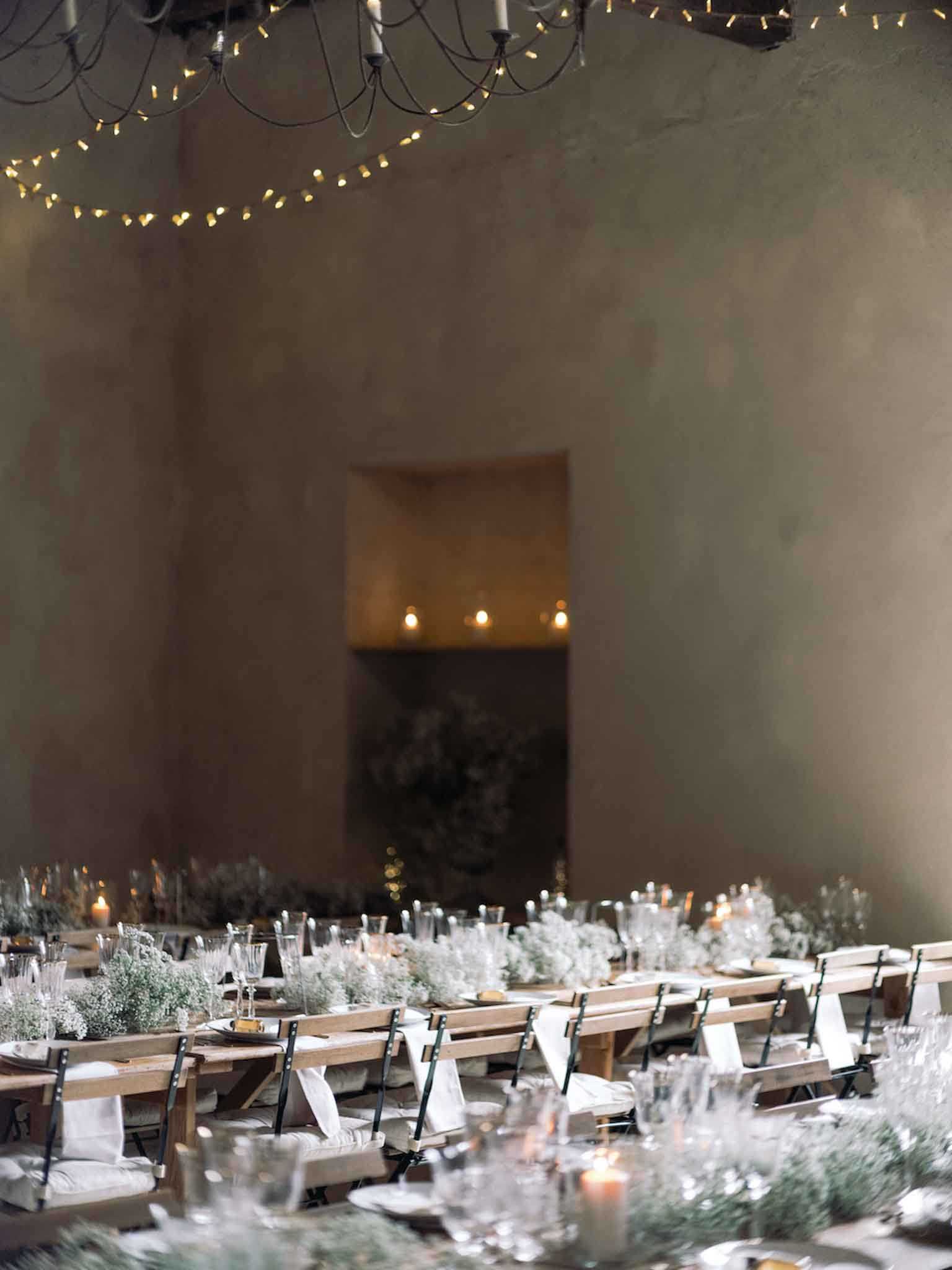 An indoor wedding reception setup photographed in a wide shot, showing long wooden farm-style trestle tables lined with black metal folding chairs with white linen seat pads and white napkins. The table runners consist of dense arrangements of baby's breath and soft grey-green foliage, interspersed with candles and clear glassware. The venue features bare plaster walls in warm taupe tones, with fairy lights strung across the ceiling alongside black wrought-iron candelabra-style chandeliers. A recessed arched niche in the background is lit with warm candlelight and filled with lush greenery, creating a focal point at the end of the room. The overall decor palette is neutral and natural — warm whites, greens, and raw wood — with a minimalist, organic styling approach. Potential venue feature image.