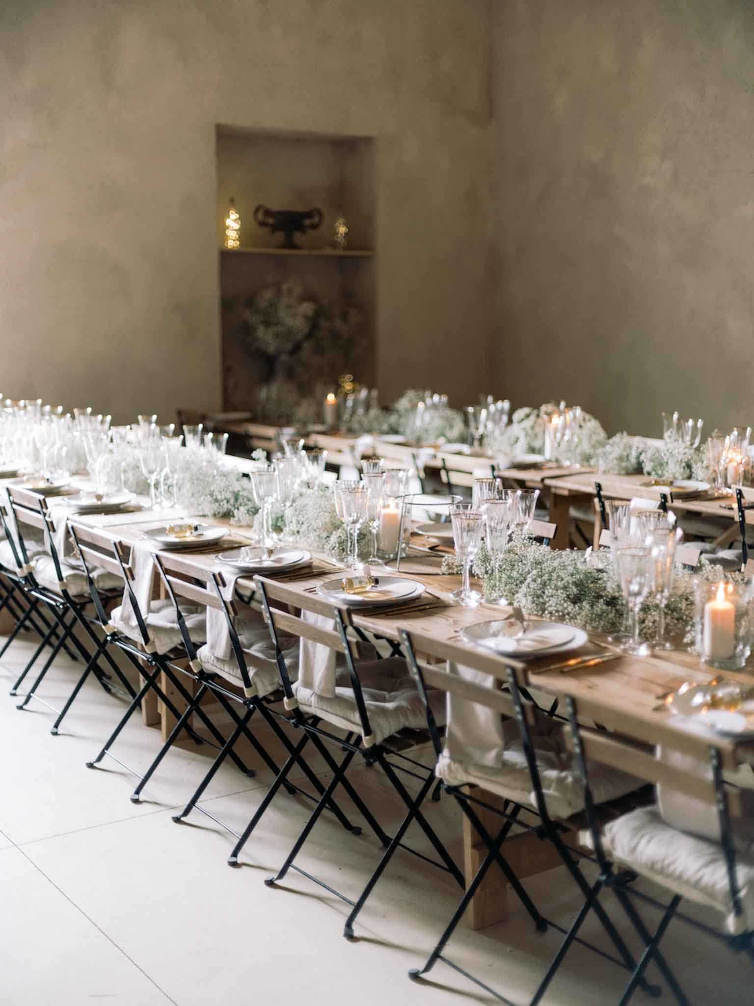 Indoor wedding reception tablescape shot from a slightly elevated angle, showing multiple long rustic wooden farm tables set for a dinner reception inside a room with bare plaster walls in a warm taupe tone. The tables are lined with black metal folding chairs with off-white linen cushions. The centerpieces consist of dense runners of white baby's breath (gypsophila) running the full length of each table, interspersed with pillar candles in glass holders and tall clear glassware including champagne flutes and water glasses. Place settings feature round white plates layered on gold charger plates with folded linen napkins. A recessed wall niche in the background holds a dark urn-style vase and small candle accents surrounded by a large loose arrangement of greenery. The overall decor palette is neutral — warm wood, white, soft gold, and black — with a minimalist, organic styling approach. Wide-angle shot capturing the full reception room setup without guests present.