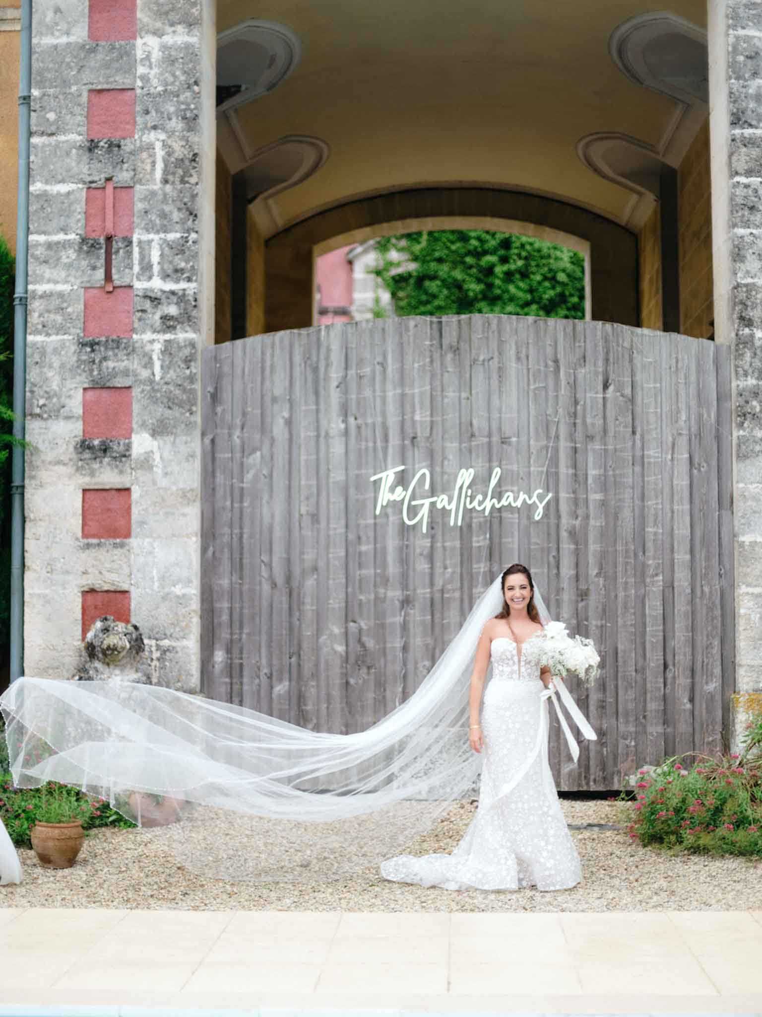 A bridal portrait taken outdoors at a French chateau or manor venue, with the bride standing on a gravel courtyard in front of a large weathered wooden gate beneath a stone archway. The bride wears a fitted, strapless lace mermaid gown with a cathedral-length veil that is billowing dramatically to the left of the frame. She holds a white bouquet of what appear to be white peonies and small white blooms tied with a white ribbon, and she is smiling directly at the camera. A white neon or acrylic script sign reading 'The Gallichans' is mounted on the wooden gate behind her. The composition is a full-length portrait shot with the architectural gateway framing the subject.