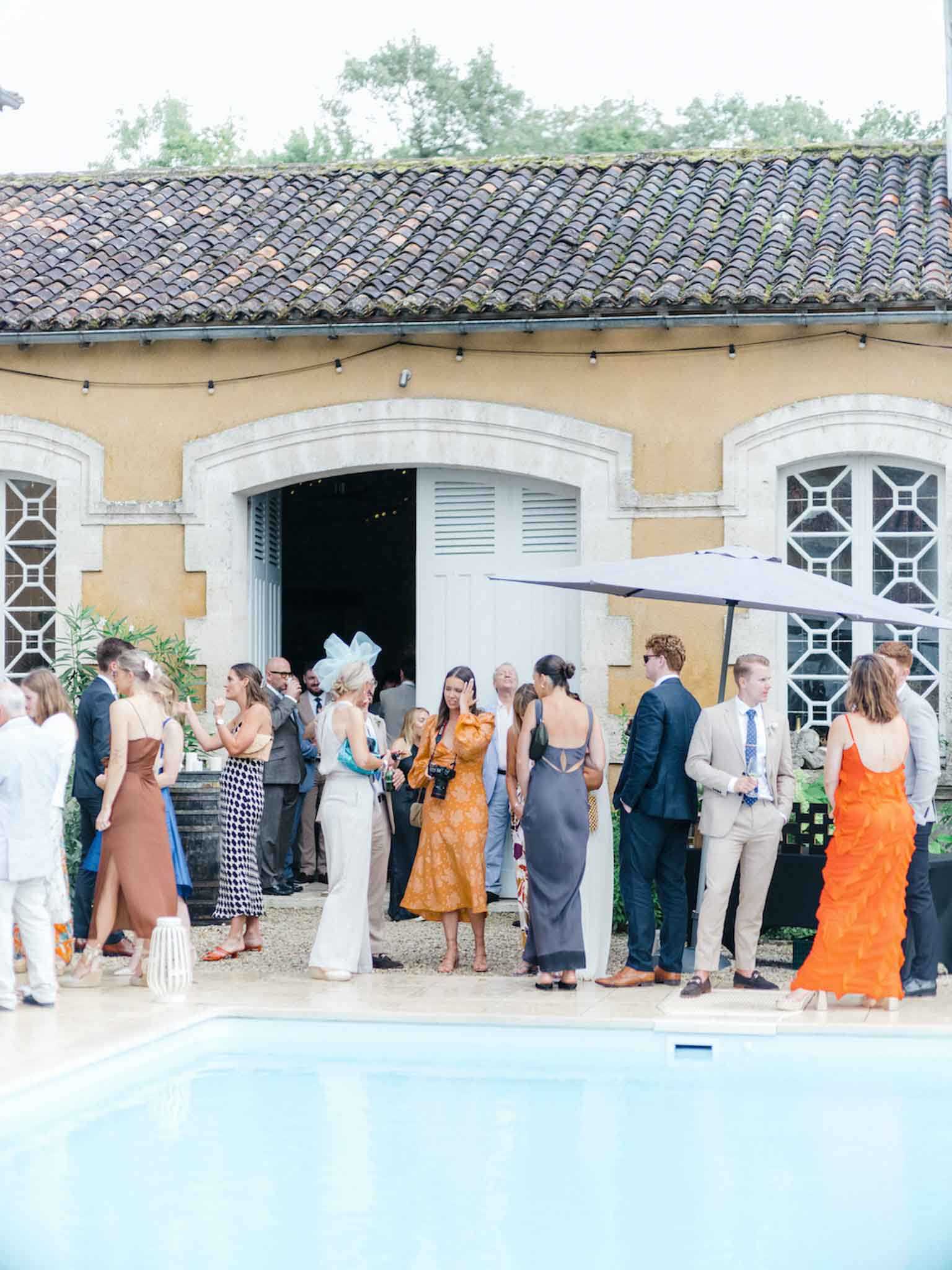 A cocktail hour is taking place outdoors at a French property, with approximately 20–25 guests mingling on a terrace alongside a swimming pool. The guests are dressed in a range of formal and semi-formal attire, including a rust-brown maxi dress, an orange ruffle gown, a mustard floral midi dress, a slate-grey slip dress, a black-and-white houndstooth outfit, and suits in navy and beige tones; one guest wears a pale blue fascinator hat. The setting features a classic ochre-rendered French outbuilding with arched stone-framed windows, louvred shutters, and terracotta roof tiles, with a dark grey patio umbrella and string lights visible. The shot is a wide, slightly distant perspective capturing the full scene of guests grouped in conversation in front of the venue building.