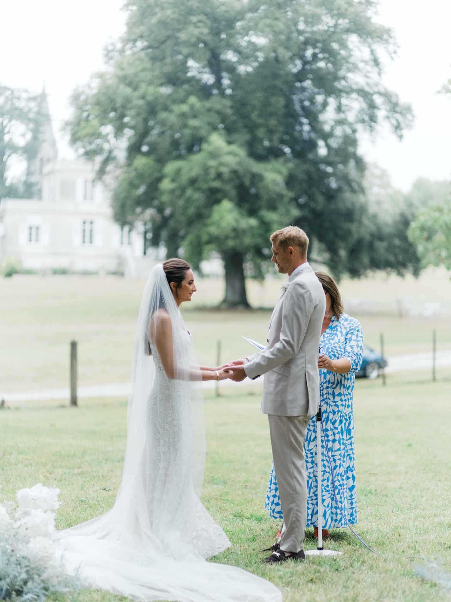 An outdoor civil or symbolic ceremony taking place on a lawn in front of a white chateau visible in the soft background. The bride wears a fitted white lace gown with a long cathedral-length veil and holds hands with the groom, who is dressed in a light grey suit; a female officiant in a blue and white patterned dress stands opposite them holding a ceremony booklet, with a microphone stand beside her. The couple faces each other in an intimate vow exchange, with white floral arrangements partially visible in the foreground. The composition is a medium full-length shot with a shallow depth of field, keeping the chateau and large trees softly blurred behind the three figures.