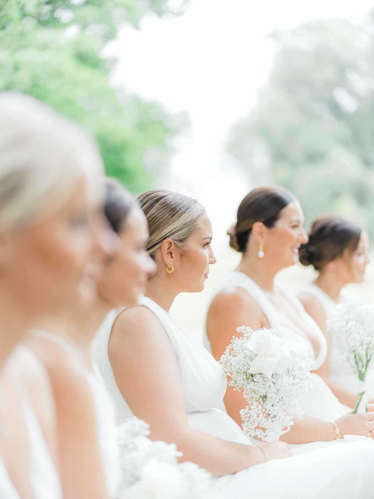 A row of approximately five bridesmaids is photographed from the side during an outdoor ceremony, all facing the same direction as they watch the proceedings. The bridesmaids wear matching white sleeveless halter-neck dresses with a clean, modern cut, and their hair is styled in sleek upswept looks including low buns and slicked-back styles. Each bridesmaid holds a bouquet composed of white baby's breath and white peonies or ranunculus, consistent across the group. The shot is a shallow-depth-of-field portrait taken from a low angle, with the nearest bridesmaid softly blurred in the foreground and focus falling on the second figure, creating a receding line effect across the frame.
