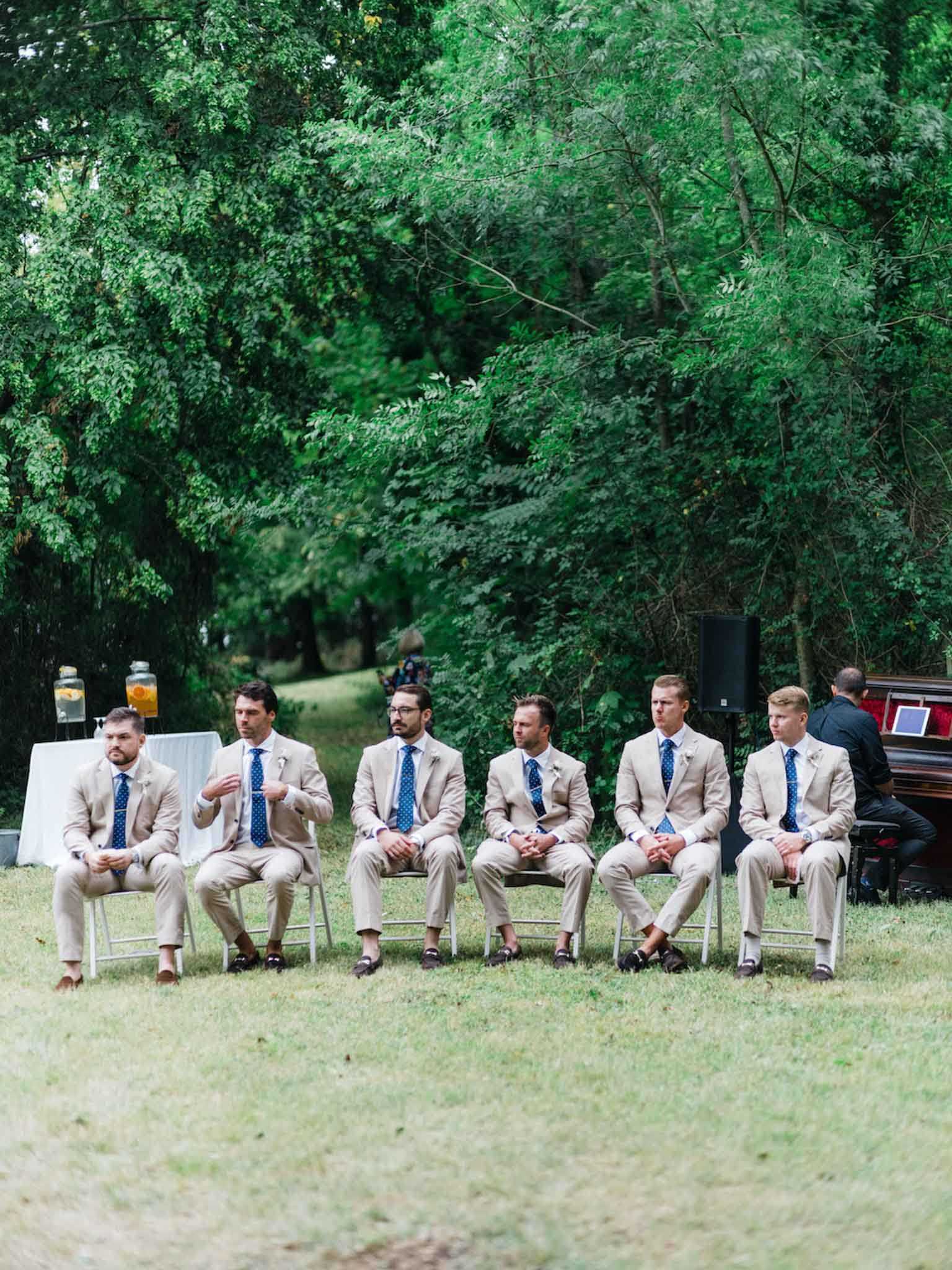 Six groomsmen are seated in a row on white folding chairs during an outdoor wedding ceremony set on a lawn surrounded by dense trees. All six men wear matching tan/sand linen suits with navy blue polka-dot ties and dark leather loafer-style shoes, each with a small white boutonniere. To the far right, a musician in black is seated at a dark upright piano, and a white linen-covered table with glass beverage dispensers is visible to the far left. The shot is a wide, eye-level portrait taken from across the lawn, capturing the full row of groomsmen as they look toward the ceremony proceedings.
