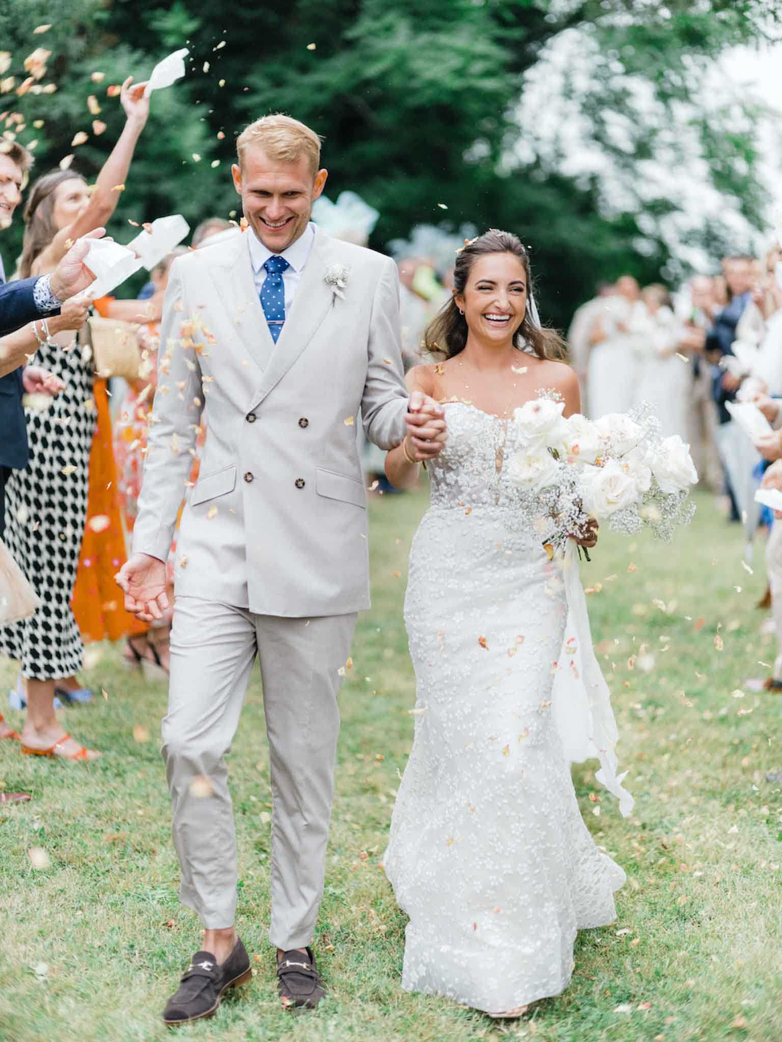 The bride and groom walk hand-in-hand through a confetti exit on an outdoor lawn, surrounded by two lines of guests tossing orange, peach, and ivory dried petal confetti. The groom wears a light grey double-breasted suit with a blue polka-dot tie and dark navy loafers, finished with a small boutonniere. The bride wears a fitted strapless lace gown with allover beaded embellishment and a chapel-length veil, carrying a bouquet of white roses and baby's breath. The shot is a medium portrait taken at eye level, with guests blurred in the background and confetti caught mid-air around the couple, both laughing openly.