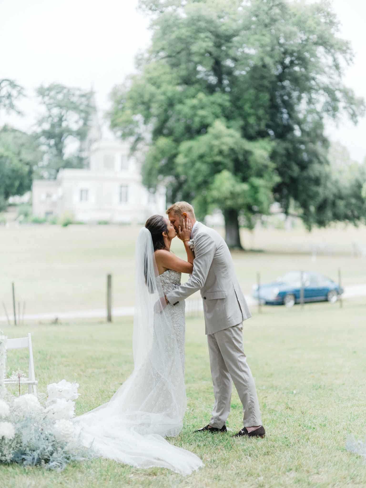 The bride and groom share a kiss during what appears to be an outdoor ceremony or couple portrait on the grounds of a French chateau, visible as a white-rendered building with a pointed tower in the soft background. The bride wears a fitted, lace-embroidered white gown with a long cathedral-length train and a shoulder-length veil, while the groom wears a light grey suit with dark loafers and a white floral boutonniere. At the lower left of the frame, white floral arrangements featuring large blooms and dusty miller are visible, likely marking the ceremony aisle. The composition is a full-length portrait shot with shallow depth of field, giving the background a soft, airy quality.