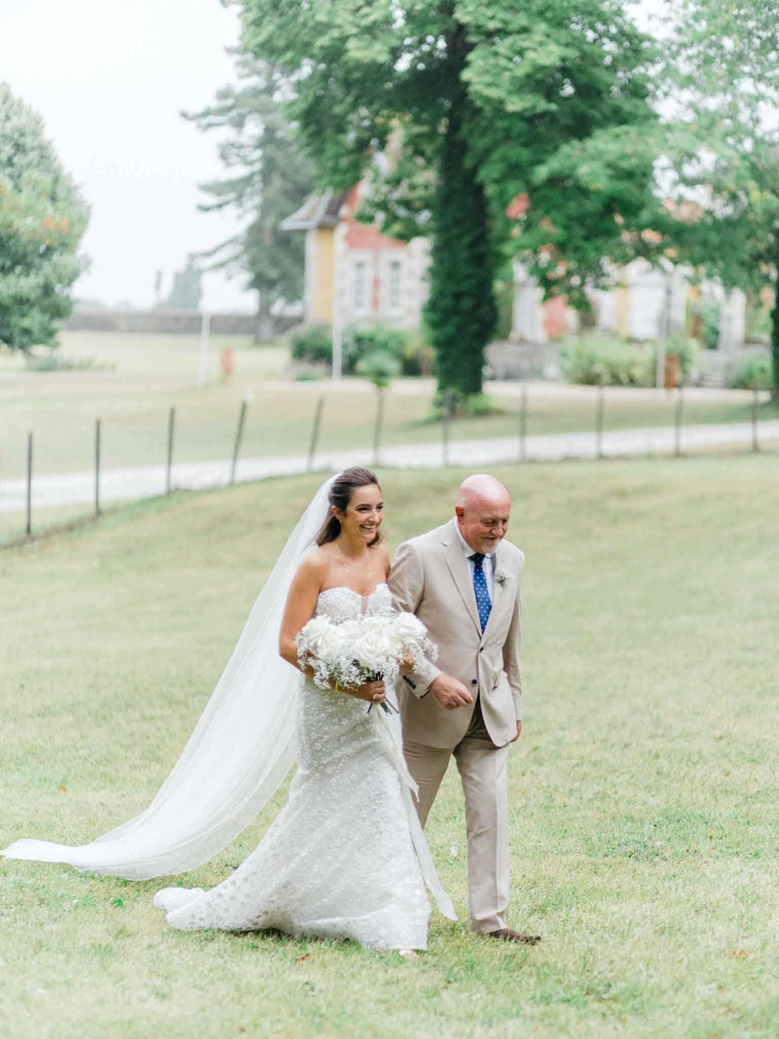 A bride and an older male companion — likely her father — walk together across a lawn toward the ceremony in an outdoor garden setting, with a chateau or manor house visible in the soft background. The bride wears a strapless, fitted lace gown with a cathedral-length veil and carries a large round bouquet of white peonies and baby's breath. She is smiling broadly, while her companion wears a tan suit with a blue polka-dot tie. The shot is a mid-length portrait taken at a slight distance, capturing the full figures and the expanse of the grounds around them.