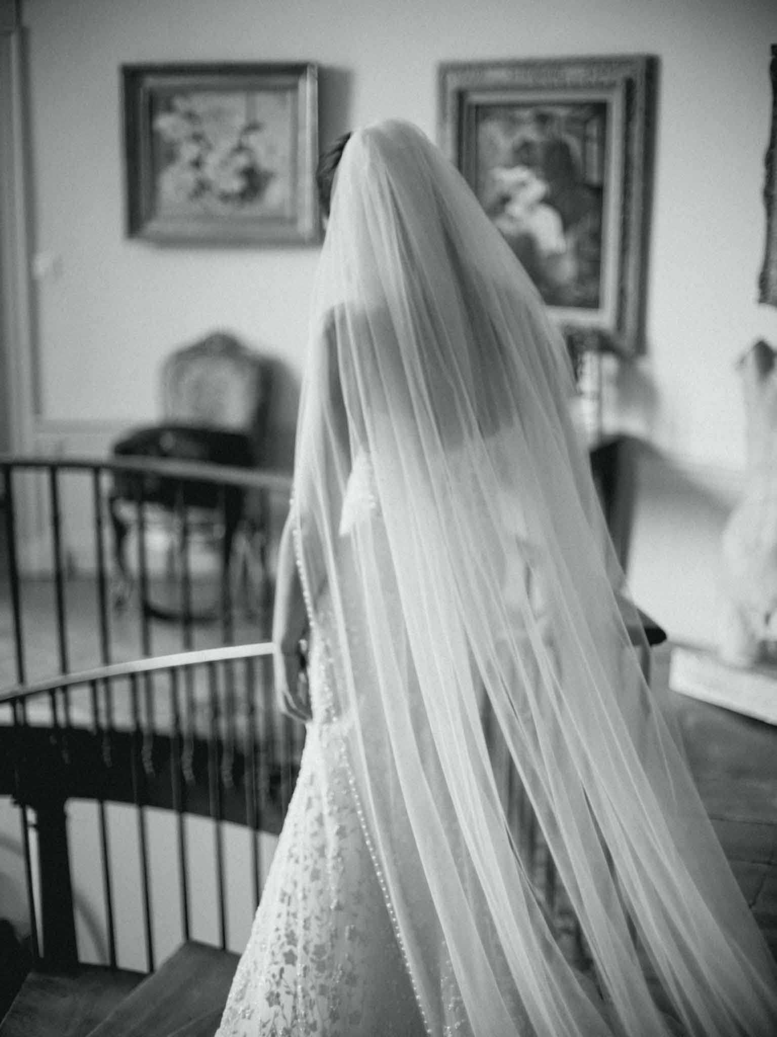 This black-and-white portrait shows a bride photographed from behind, standing at the top of a curved staircase with a wrought iron railing inside what appears to be a chateau or manor house. She is wearing a lace wedding gown with floral appliqué detailing and a long cathedral-length veil with a delicate beaded or pearl edge that cascades down her back. The interior setting features ornate gilt-framed paintings on the walls and antique furniture visible in the soft background. The image has soft contrast with bright highlights on the veil fabric, and is composed as a close-to-medium portrait focused on the veil and gown detail.