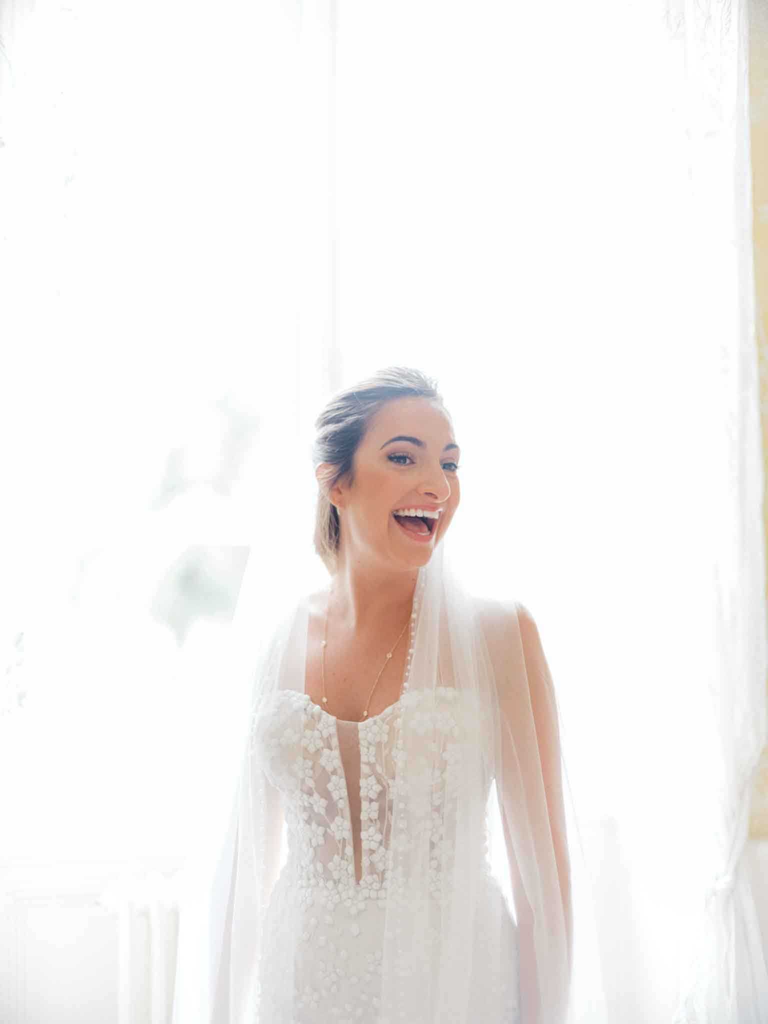 A bridal portrait taken indoors, likely during the getting-ready phase, with the bride positioned in front of a large window with sheer white curtains creating a bright, backlit effect. The bride is laughing openly, wearing an ivory fitted gown with a deep V-neckline and 3D floral appliqué detailing across the bodice, paired with a long plain-edge tulle veil draped over her shoulders and a delicate layered pendant necklace. Her dark hair is pulled back into a low updo. The composition is a medium portrait shot, framing the bride from approximately the waist up against the overexposed window light.