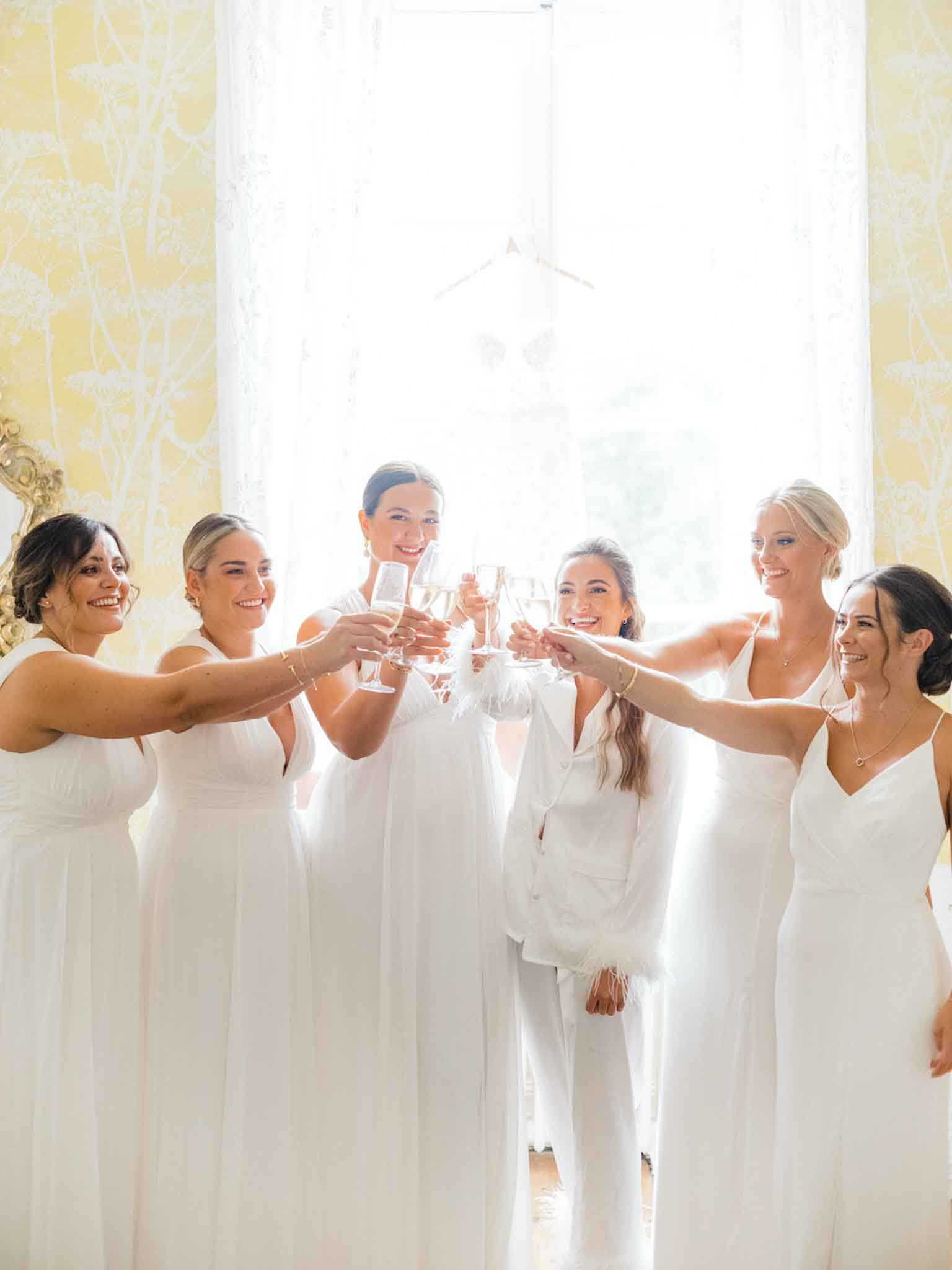 A getting-ready moment showing a bride and five bridesmaids toasting with champagne flutes inside what appears to be a chateau or manor house bridal suite. The bride is dressed in a white satin pajama set with feather-trimmed cuffs, while the five bridesmaids all wear floor-length white dresses in varying styles including halter neck, sleeveless, and thin-strap silhouettes. The room features pale yellow botanical-print wallpaper and sheer white curtains over a large window, which backlight the group with soft natural light. The shot is a mid-length group portrait with all six women smiling and extending their glasses toward the center.