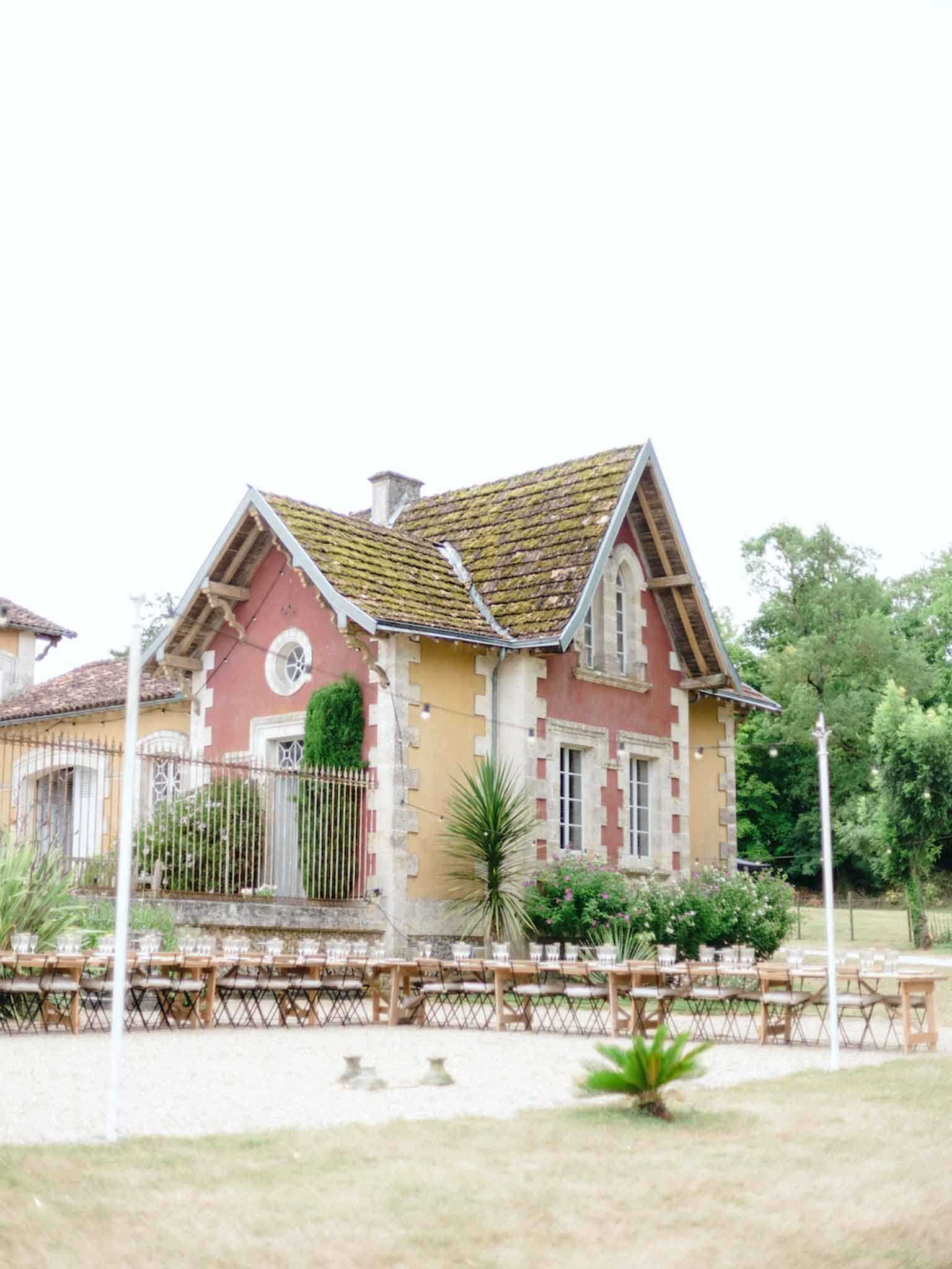 An outdoor reception setup is arranged in front of a French country estate building featuring terracotta-red and ochre rendered walls, stone quoins, arched windows, and a moss-covered pitched tile roof. Long wooden trestle tables with folding wooden chairs are arranged in a U-shape or extended rows across the gravel courtyard, set with white linens or place settings visible along the table lengths. The wide shot captures the full façade of the gatehouse-style building alongside the adjacent main property, with an ornate iron gate and climbing greenery framing the entrance. No people are present in the image, suggesting this was taken during setup before guests arrived. Potential venue feature image.