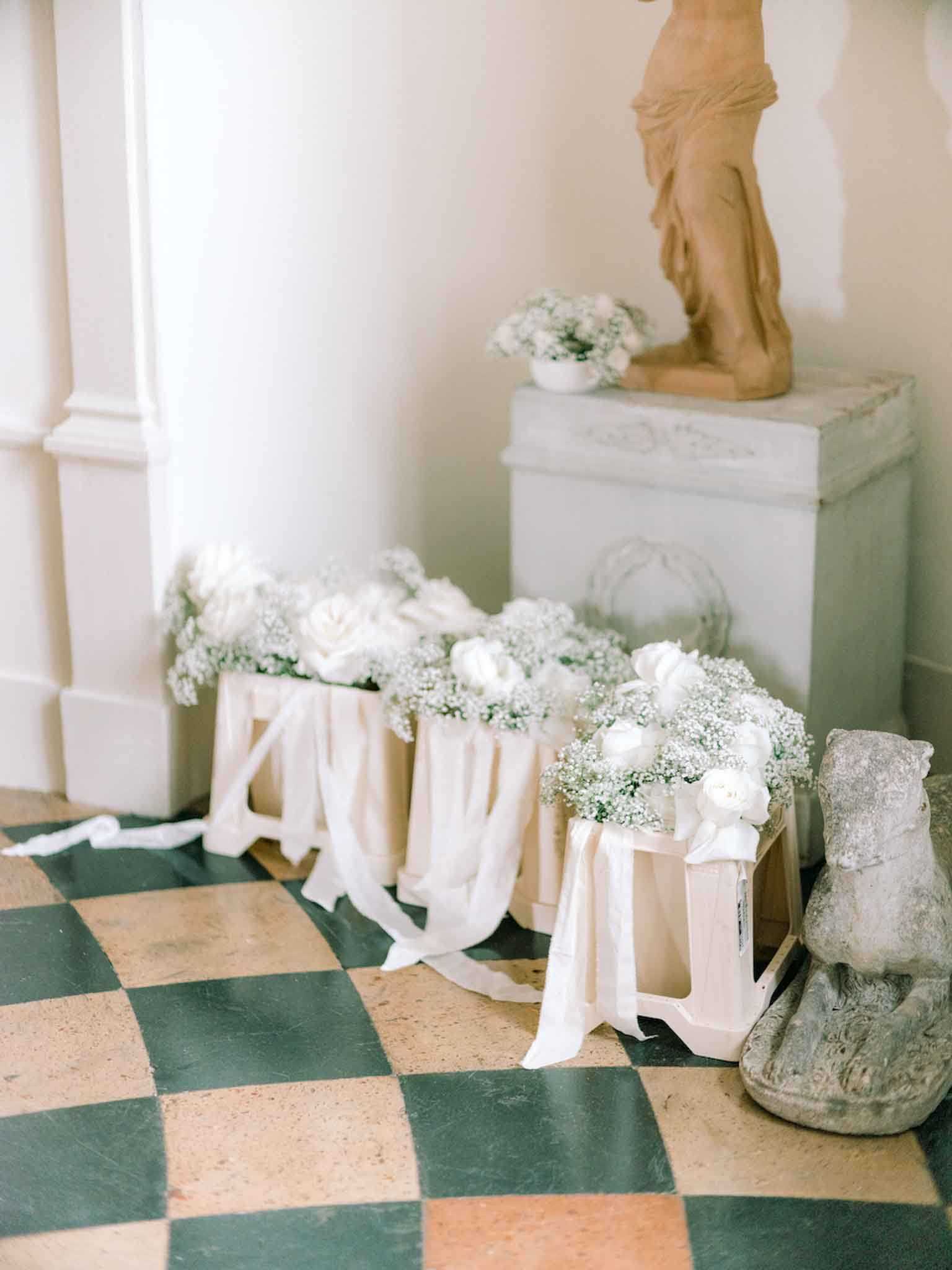 A floral detail shot taken indoors on a green and cream checkered marble floor, showing three small white wooden stools or crates lined up against a wall, each topped with arrangements of white baby's breath and white garden roses, with cream silk or chiffon ribbon draped loosely over each one and trailing onto the floor. Behind the stools sits a white painted stone pedestal holding a bowl of matching white baby's breath, topped with a classical terracotta figurine sculpture. A weathered stone decorative fragment is visible on the right side. The overall decor palette is all-white and cream with a classic French interior aesthetic. Close-up detail composition.