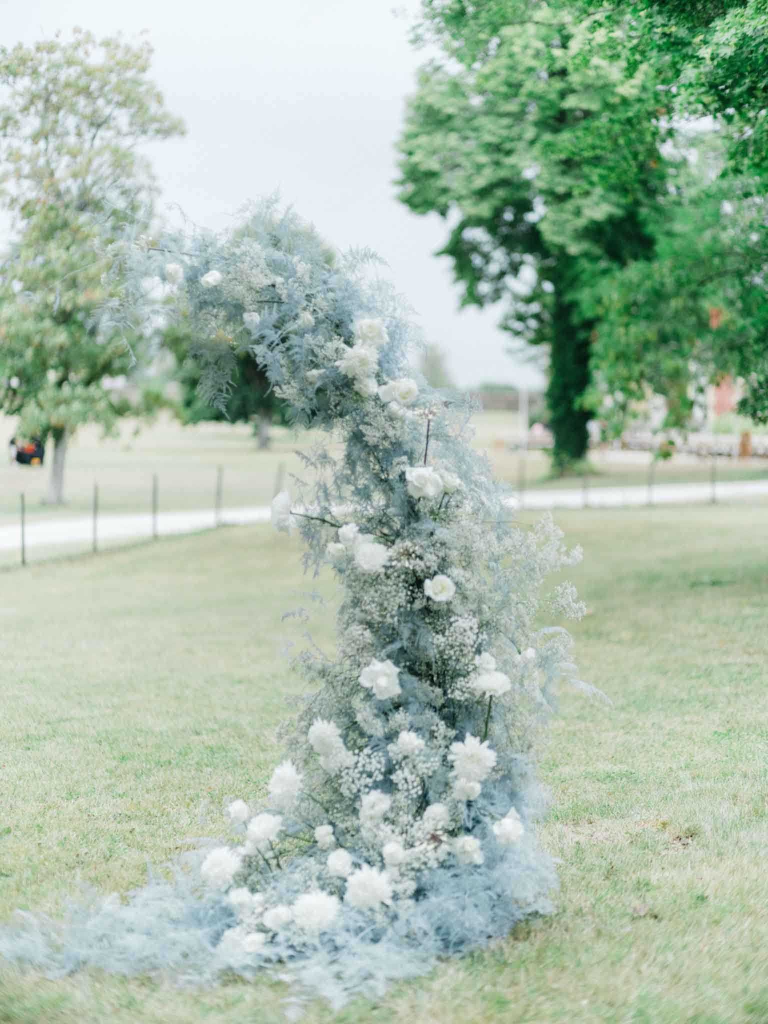 A close-up detail shot of a freestanding floral column arrangement placed on a lawn at an outdoor wedding ceremony. The arrangement is composed of white garden roses, white carnations, baby's breath, and abundant dusty blue-grey foliage — likely dried or dyed asparagus fern and similar soft, feathery greenery — creating an airy, tonal palette of white and muted blue-grey. The column rises from the grass and curves slightly at the top, with flowers and foliage spilling onto the ground at the base. The overall floral styling is organic and loosely structured, consistent with a romantic, soft classical aesthetic. The background is softly blurred, revealing an open parkland setting with trees and a fence line in the distance.