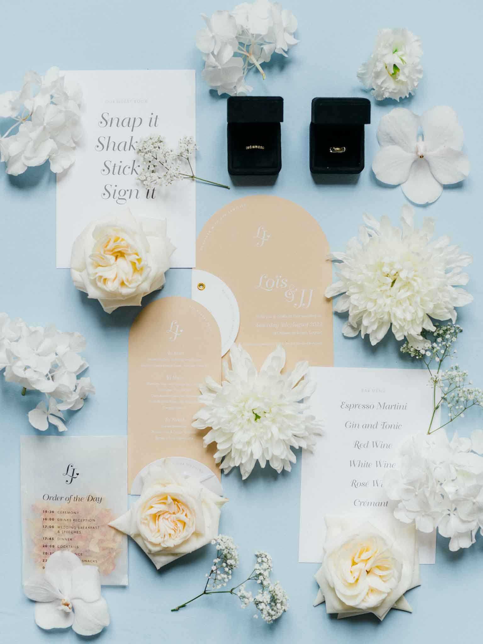 A flat lay detail shot of a wedding stationery suite and accessories arranged on a pale blue background. The stationery set includes arch-shaped invitations in warm sand/tan with white typography bearing the names 'Lois & JJ,' an order of the day card on translucent vellum, a drinks menu listing espresso martini, gin and tonic, red wine, white wine, rosé wine, and crémant, and a guest book instruction card reading 'Snap it, Shake it, Stick it, Sign it.' Two gold wedding bands are displayed in open black velvet ring boxes at the top of the frame. The styling palette is minimal and modern, using clean arch shapes and serif typography in sand and white. Scattered throughout the composition are cream garden roses, white dahlias, white hydrangea petals, and sprigs of baby's breath, all in white and soft cream tones.
