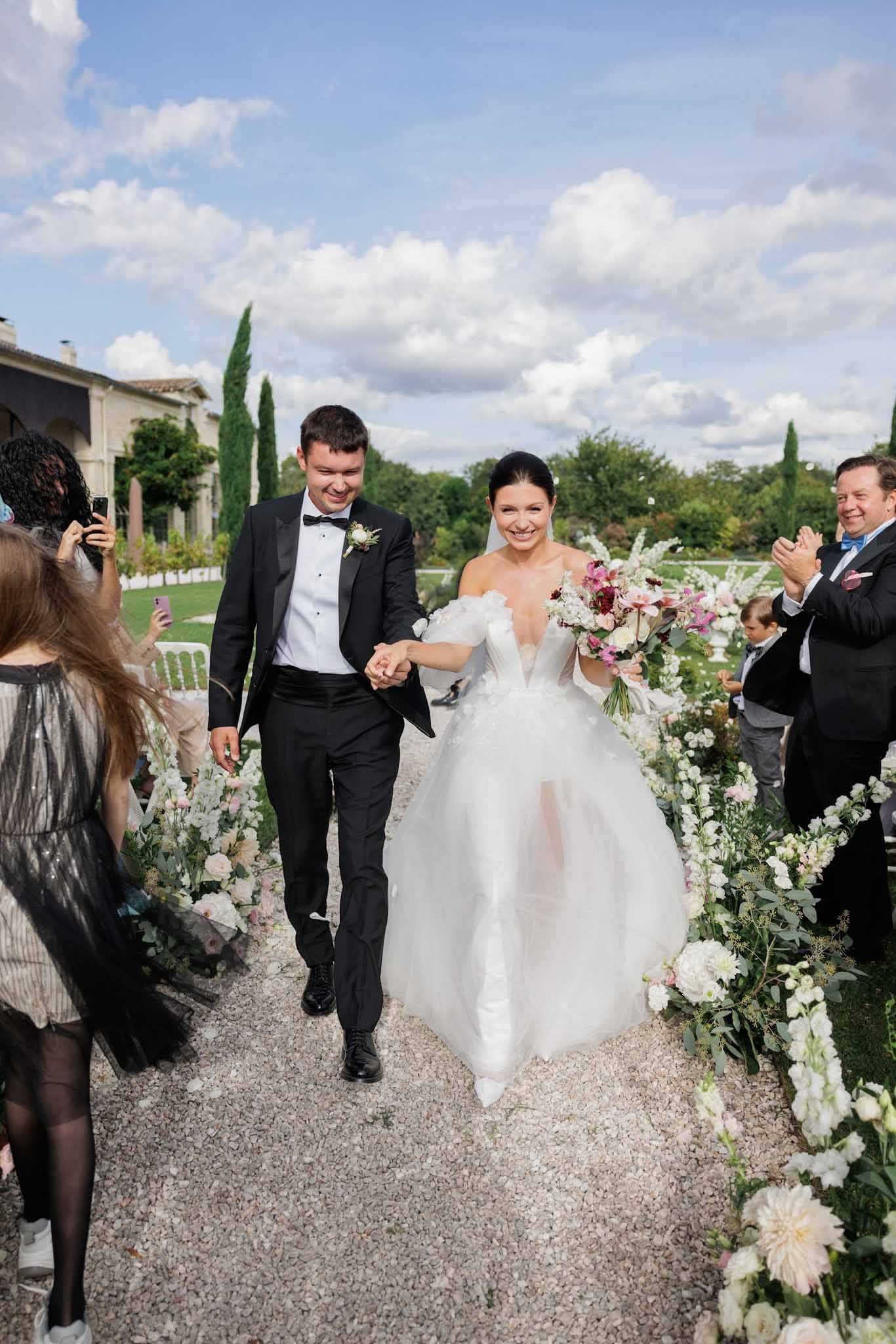 The couple is walking back up the aisle as newlyweds following an outdoor ceremony at what appears to be a French chateau, with the stone building and tall cypress trees visible in the background. The bride wears a full-ball-gown white dress with a deep V-neckline, off-the-shoulder ruffle sleeves, and white pointed-toe heels, carrying a lush bouquet featuring deep burgundy, pink, and white blooms including what appear to be orchids, garden roses, and astilbe. The groom is dressed in a black tuxedo with a black bow tie and a small floral boutonniere. The gravel aisle is flanked by low floral arrangements in white, blush pink, and cream tones with eucalyptus greenery, and guests on both sides are clapping and photographing the couple; the overall decor palette is classic black and white with romantic blush and burgundy floral accents.