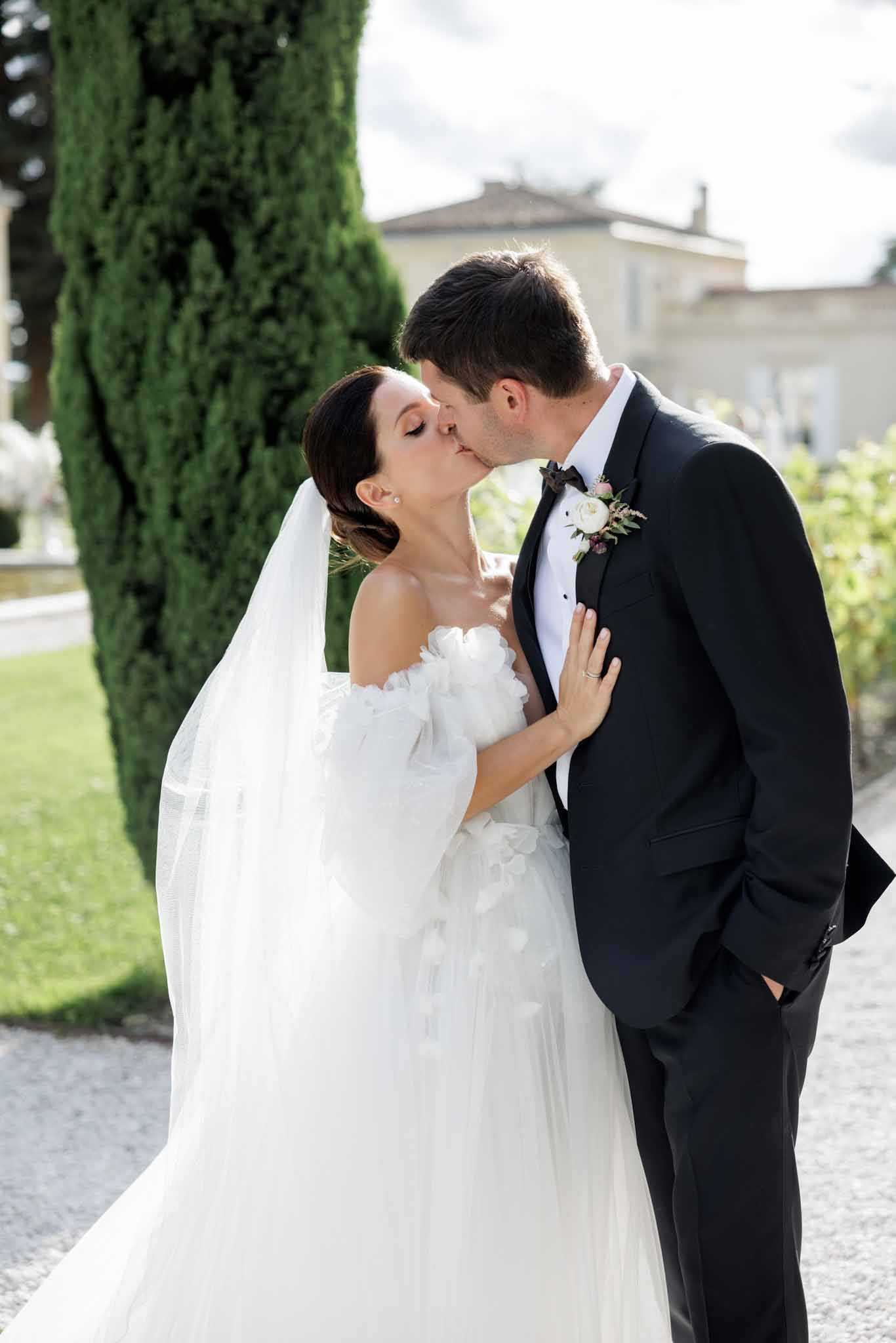 A couple portrait shot outdoors on a gravel path, with the bride and groom sharing a kiss. The bride wears an off-the-shoulder white tulle ball gown with three-dimensional floral appliqué detailing and a long flowing veil, her dark hair styled in an updo. The groom wears a classic black tuxedo with a white dress shirt and black bow tie, with a boutonnière featuring a white rose, small pink flowers, and greenery. The setting appears to be the grounds of a French chateau or manor house, with a tall cypress tree and a stone building visible in the soft background. The composition is a mid-length portrait with a shallow depth of field, keeping the couple sharply in focus against the blurred garden setting.