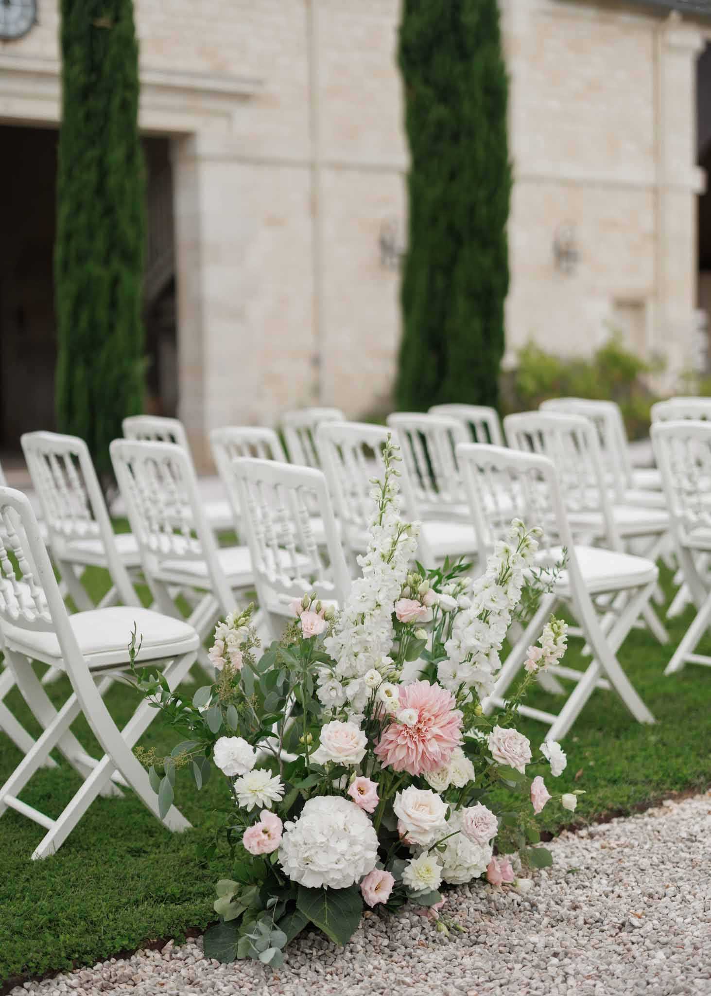 An outdoor wedding ceremony setup on the grounds of a French chateau, with multiple rows of white Napoleon-style chairs arranged on a grass lawn beside a gravel aisle. In the foreground, a close-up aisle floral arrangement features white hydrangeas, blush pink dahlias, light pink roses, white delphiniums, blush lisianthus, and eucalyptus foliage. The background shows the limestone facade of the chateau with tall Italian cypress trees framing the entrance, contributing to a classic French formal garden aesthetic. The composition is a detail/portrait-orientation shot with shallow depth of field, keeping the floral arrangement sharp while the chairs and building recede softly out of focus. Potential venue feature image.