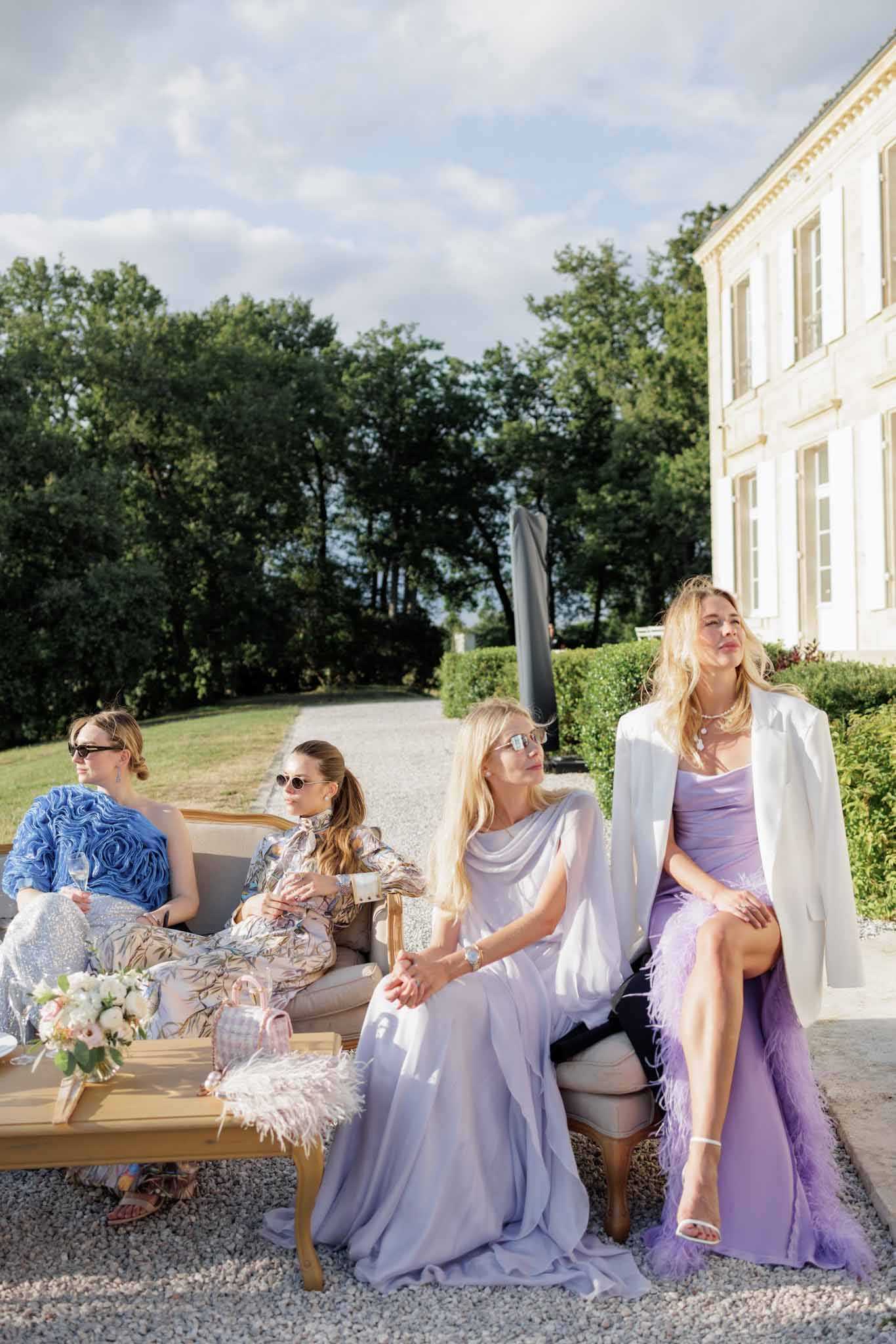 Four female guests are seated on gold Louis XVI-style chairs and a low wooden coffee table during what appears to be an outdoor cocktail hour on the gravel grounds of a French chateau. The white classical chateau building is visible in the right background. The women wear distinctly styled outfits: one in a blue textured ruffled skirt, one in a floral printed long-sleeve blouse and trousers, one in a pale lavender draped floor-length gown with a white cape or shawl, and one in a lilac feather-trimmed dress with a white blazer. All four women wear sunglasses and appear relaxed, looking away from the camera. A small bouquet of blush and ivory flowers with greenery sits on the coffee table alongside what appears to be a pink gingham bag. The overall styling palette is soft lavender, lilac, and pastel tones with feather and textured fabric details suggesting a fashion-forward, modern-chic aesthetic. Medium wide shot, taken at ground level in natural afternoon light.
