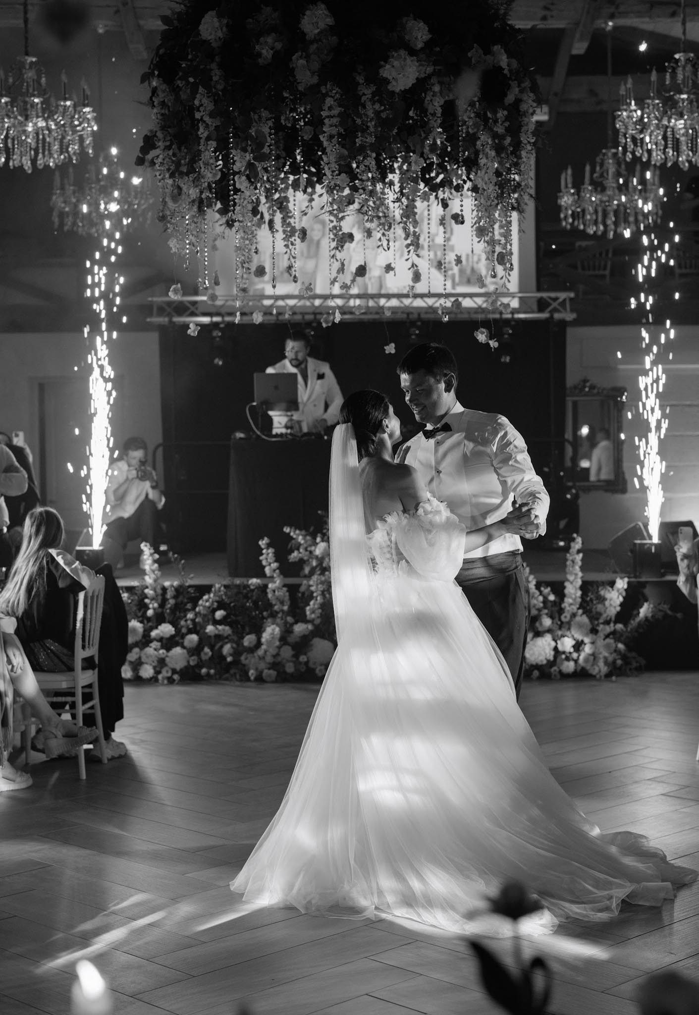 This black-and-white image captures a couple's first dance in an indoor ballroom with a herringbone parquet floor. The bride wears a full tulle skirt gown with a long veil and holds a small bouquet, while the groom is dressed in a white dress shirt and black bow tie; the two face each other closely and smile. Cold sparkler fountains flare on either side of the dance floor behind them, with a DJ visible at a booth in the background and large floral ground arrangements flanking the stage area. Overhead, a suspended floral installation with trailing greenery and crystal chandelier accents frames the scene, and several guests seated around the perimeter watch the dance. The image is a medium-wide portrait shot with high contrast, the bright tulle of the bride's dress standing out sharply against the darker surroundings.