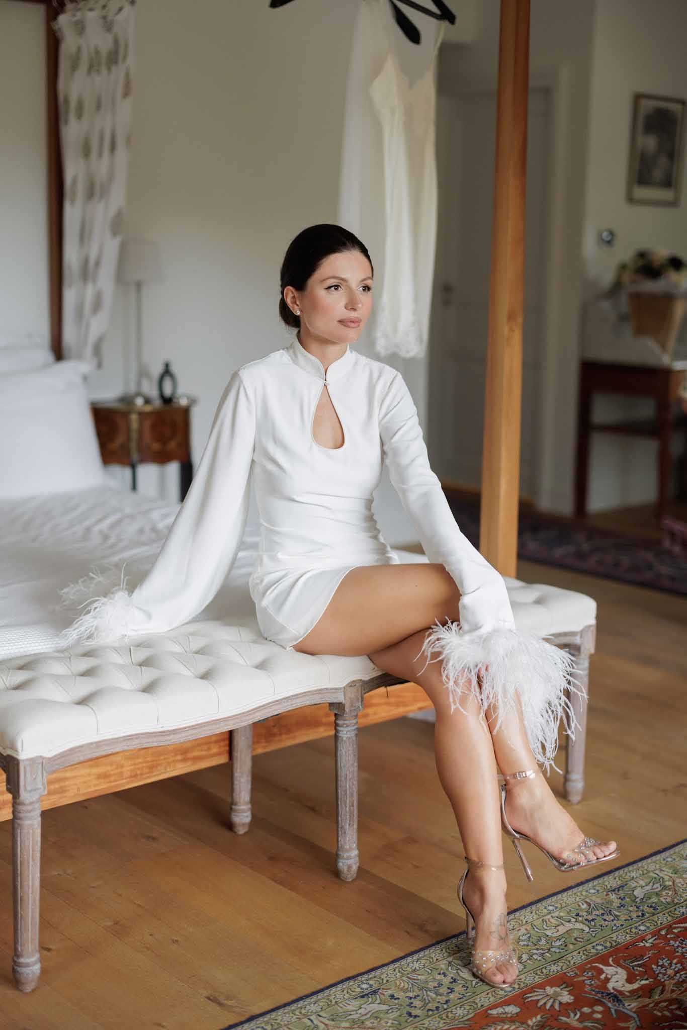 A getting-ready portrait of a bride seated on a tufted white upholstered bench with grey-washed wooden legs, positioned in what appears to be a chateau bedroom with hardwood floors and a patterned area rug. The bride wears a short white mini dress featuring a mandarin collar, a keyhole cutout at the chest, long sleeves with white feather trim at the cuffs, and pairs it with strappy clear crystal stiletto heels. Her dark hair is pulled back in a sleek low bun and she gazes off to the side. In the background, a full-length white bridal gown is visible hanging on a wooden beam, suggesting a dress change is planned, and the room is furnished with antique-style wooden pieces. The composition is a full-length portrait shot with a modern, fashion-forward bridal styling aesthetic.