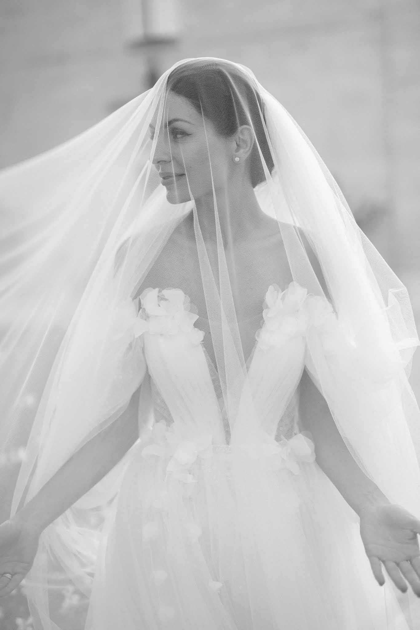 A black-and-white bridal portrait showing a bride from approximately the waist up, photographed outdoors against a softly blurred architectural background. The bride wears a voluminous white ball gown with a deep V-neckline and three-dimensional floral appliqués scattered across the bodice and shoulders, styled in a classic formal aesthetic. A long, flowing cathedral-length veil is draped over her head and spreads wide around her, its sheer tulle fabric catching light and creating strong contrast against the bright tones of her gown. The bride faces slightly to the left in a composed profile pose, wearing small stud earrings, with her hair neatly pulled back, and a subtle smile on her face.