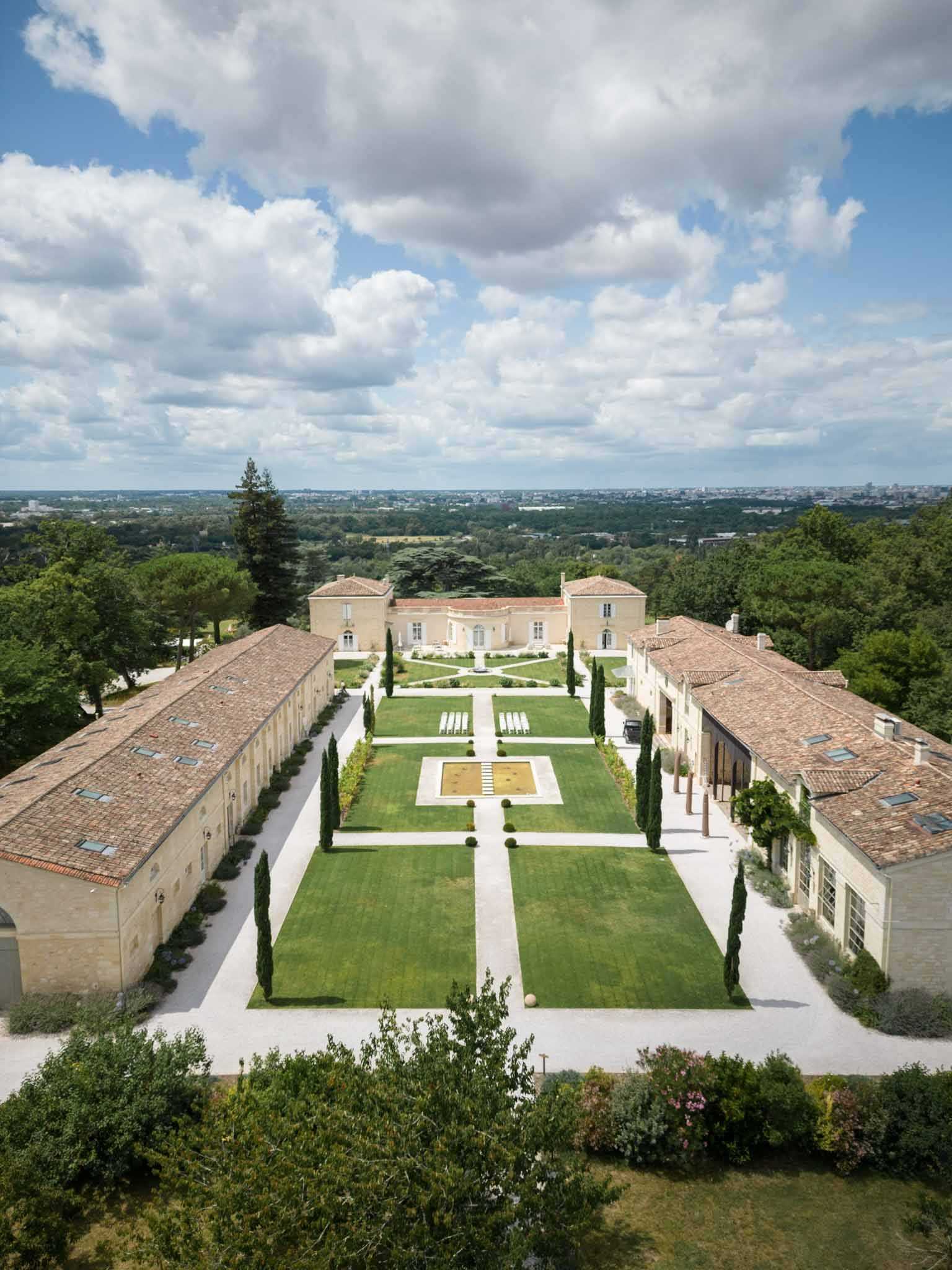An aerial wide shot of a French chateau estate and its formal gardens, with no wedding guests visible. The property features a symmetrical French formal garden layout with clipped cypress trees lining a central axis, a rectangular reflecting pool or water feature at the center, geometrically divided lawn panels, and white gravel pathways. A row of white ceremony chairs is set up on the lawn mid-ground, indicating a ceremony space in preparation. The main chateau building in warm limestone with terracotta roof tiles sits at the far end of the garden, flanked by two long stone outbuilding wings with matching terracotta roofs on either side. The surrounding landscape shows an expansive wooded area with a city skyline visible in the far distance. Potential venue feature image.