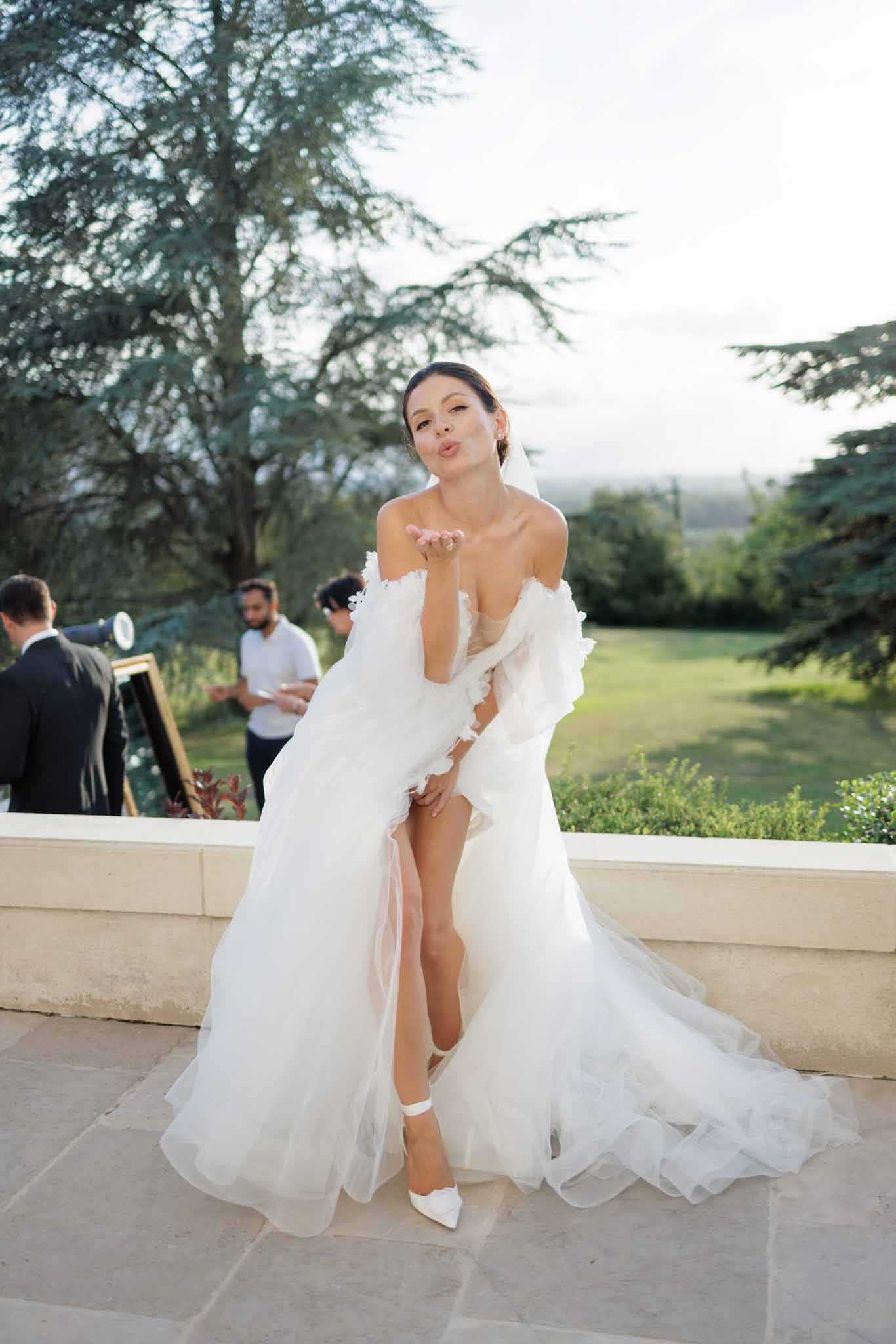 A bride poses playfully on an outdoor stone terrace, blowing a kiss toward the camera with one hand raised to her lips. She wears an off-the-shoulder white tulle ball gown with ruffled detailing along the sleeves and bodice, a high front slit, a cathedral-length train, and white pointed-toe heels with ankle straps; a sheer veil is visible at the back of her upswept hair. Two guests or vendors are visible in the soft background, one near a gold-framed easel, with an open landscaped garden and rolling countryside beyond the terrace balustrade. The shot is a full-length portrait with a shallow depth of field, giving the background a blurred quality while keeping the bride in sharp focus.