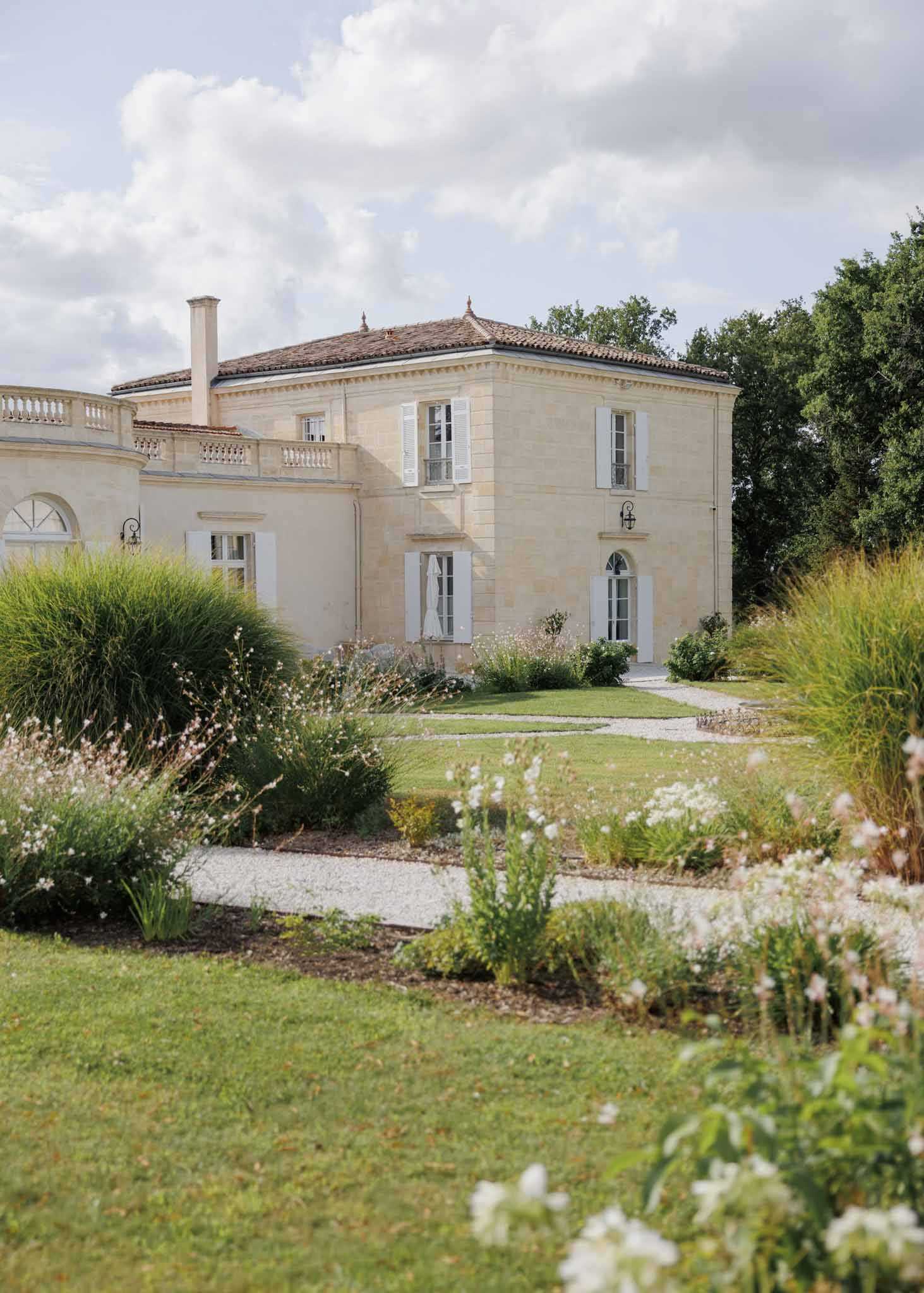 Wide exterior shot of a French château-style manor house featuring cream limestone façade, white shuttered windows, terracotta tiled roof, and a curved balustraded wing visible on the left side. A gravel pathway leads toward the entrance, flanked by landscaped garden beds with ornamental grasses, white and pale pink wildflowers, and flowering shrubs in the foreground. No people are visible in the image. Potential venue feature image.