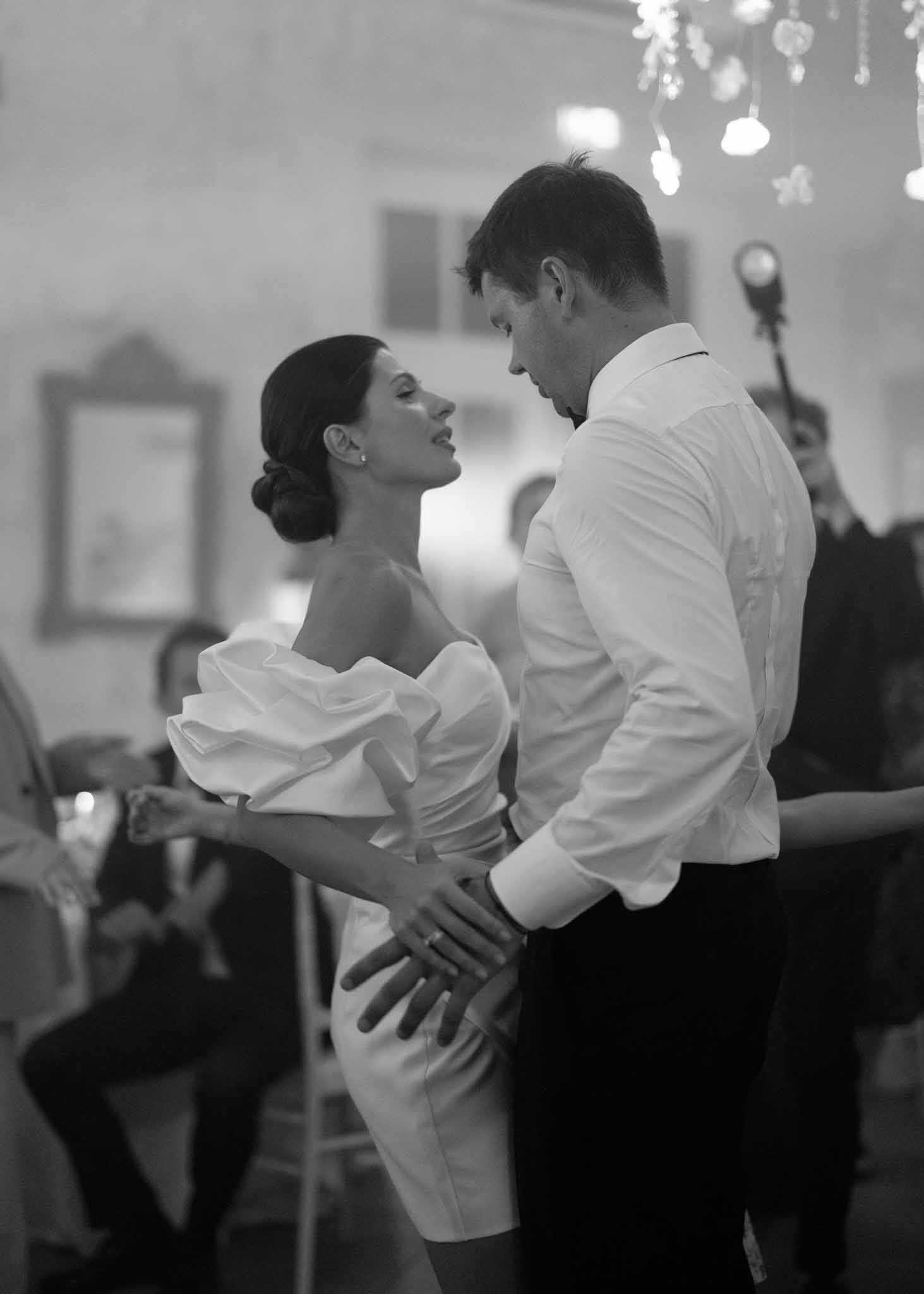 This black-and-white image captures a couple's first dance at an indoor reception, likely inside a chateau or historic ballroom. The bride wears a short, structured off-the-shoulder dress with dramatic ruffled sleeves and a fitted silhouette, her dark hair styled in a low chignon with pearl earrings; the groom is in a white dress shirt and dark trousers, his jacket removed. The two face each other closely, his hands on her waist and hers on his, with guests visible and slightly blurred in the background watching the moment. A chandelier with floral or crystal detailing is partially visible at the top of the frame, and framed artwork hangs on the light-toned walls, suggesting a classic, formal interior setting; the image is a medium portrait shot with shallow depth of field, rendering the background in soft, low-contrast tones.