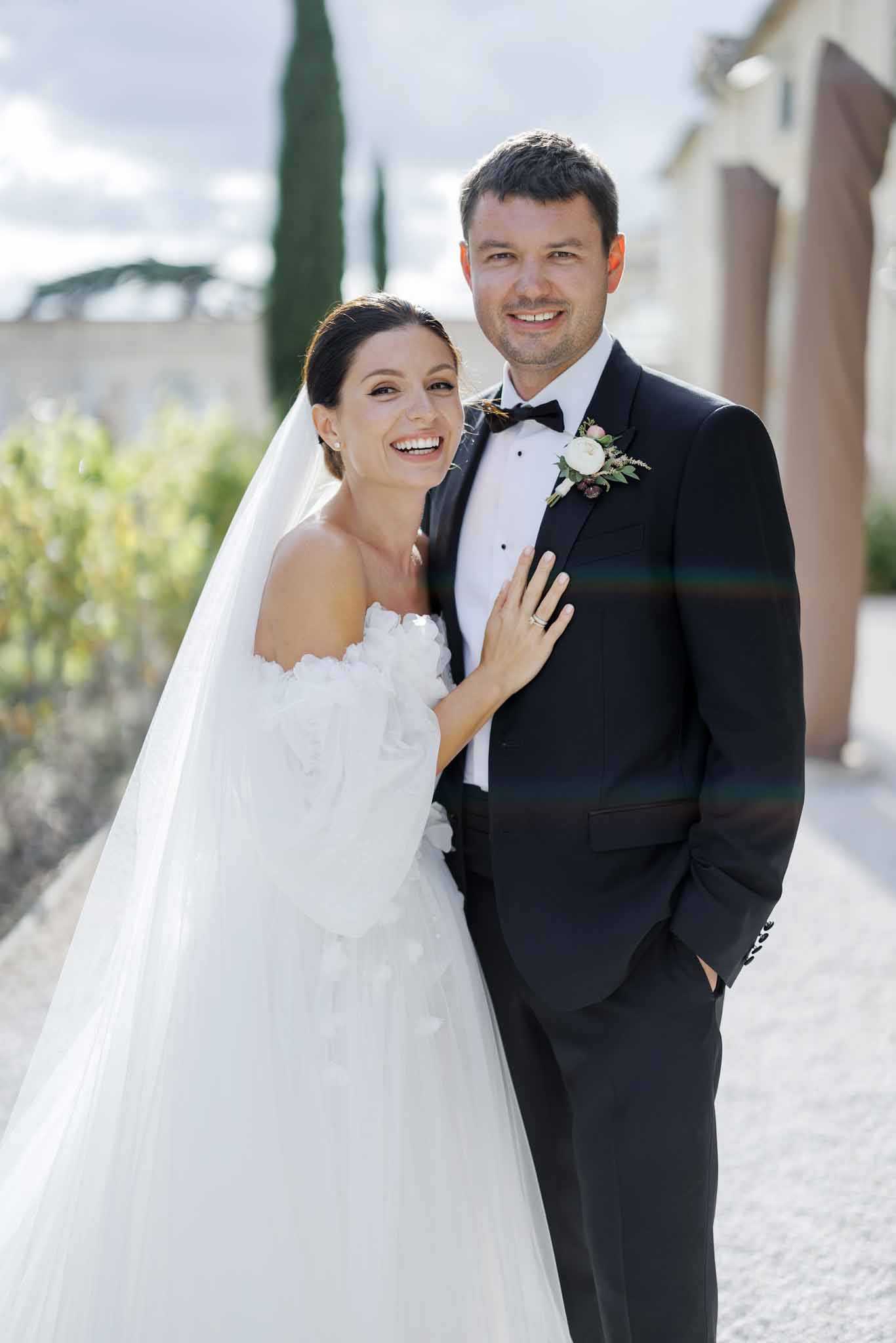 A couple portrait taken outdoors on a gravel path in front of a Provençal-style building with warm ochre tones. The bride wears an off-the-shoulder white ballgown with three-dimensional floral appliqué detailing and a long cathedral-length veil, with her dark hair pulled back; she rests her hand on the groom's chest and smiles broadly at the camera. The groom wears a black tuxedo with a black bow tie and a white ranunculus and greenery boutonniere with small burgundy berry accents. The shot is a mid-length couple portrait with natural daylight and a slight lens flare visible across the frame.