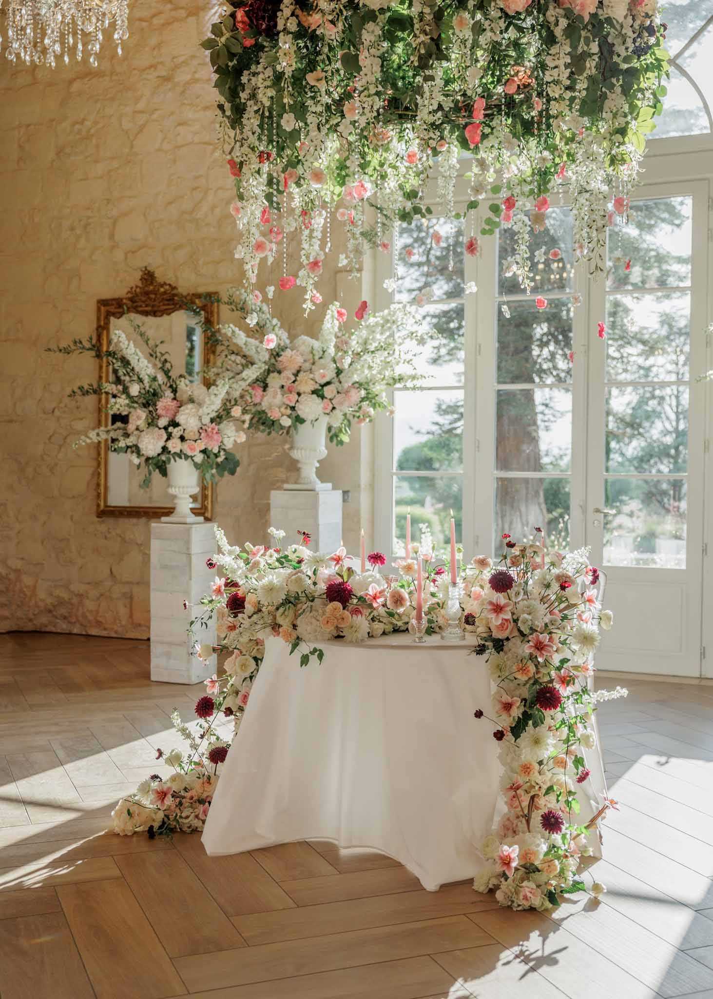 A detail shot of a sweetheart table setup inside a French chateau-style reception room with exposed limestone walls, herringbone parquet flooring, and large French doors letting in natural light. The white linen-draped table is heavily adorned with cascading floral arrangements that spill from the tabletop down the legs to the floor, featuring ivory garden roses, blush and coral dahlias, burgundy dahlias, white cosmos, and greenery. Slim blush taper candles in glass holders are placed along the center of the table. Above, a large hanging floral chandelier installation drips with white wisteria-style blooms, coral and pink roses, green foliage, and crystal strands. In the background, two large white urn pedestals hold oversized arrangements of white hydrangeas, blush peonies, and tall white flower stems, positioned in front of a gilt-framed ornate mirror. The overall decor palette is white, blush, coral, and burgundy with a romantic garden-inspired style. Potential venue feature image.