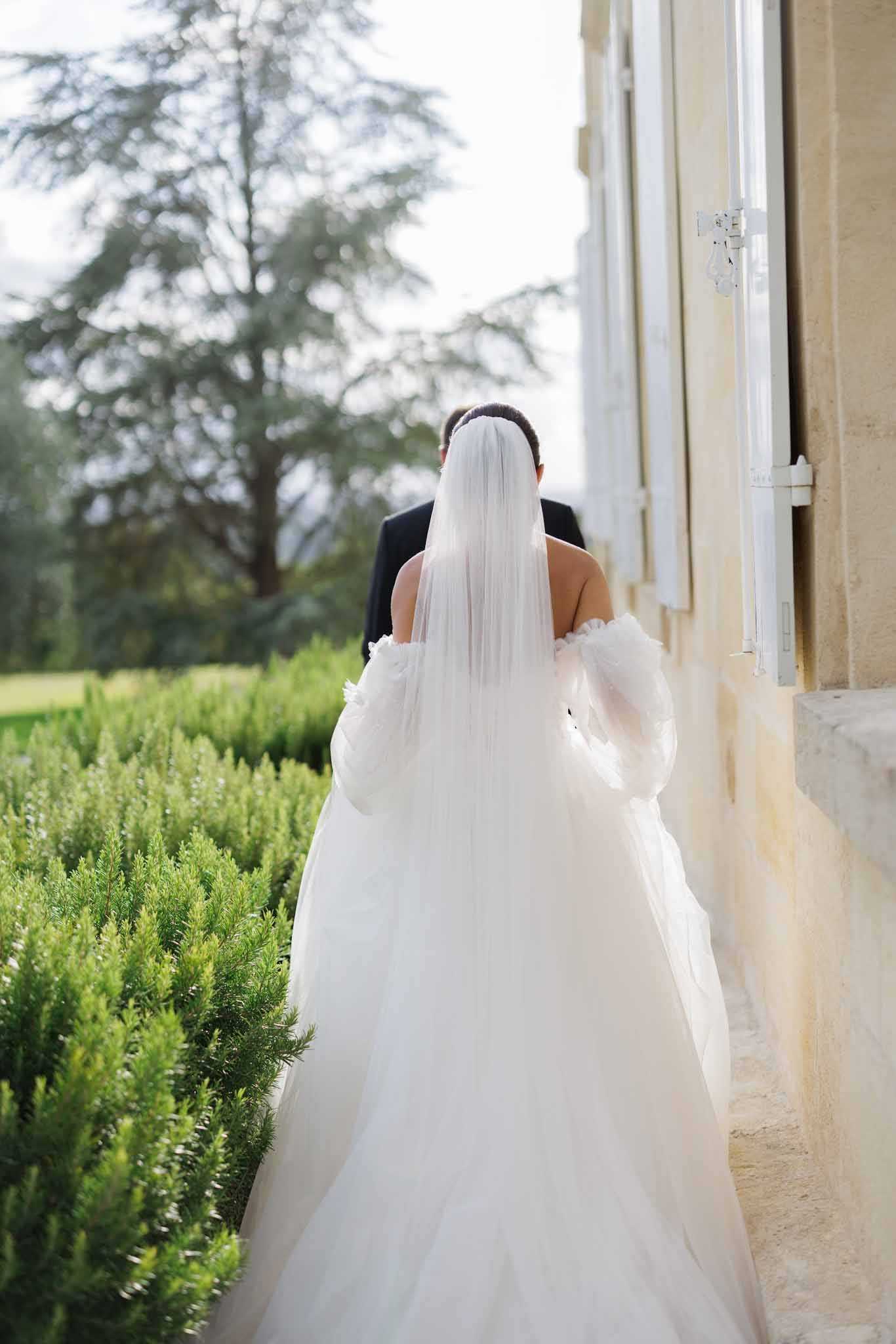 A couple walks side by side along the exterior of a chateau, photographed from behind in a medium portrait shot. The bride wears a full-volume white tulle ball gown with detachable puff sleeves and a long straight-edge veil that falls to mid-back, while the groom is dressed in a dark navy or black suit. The setting is outdoor along a formal garden terrace, with trimmed hedging running alongside the pale stone chateau facade fitted with white-shuttered windows. The overall styling is classic and formal.