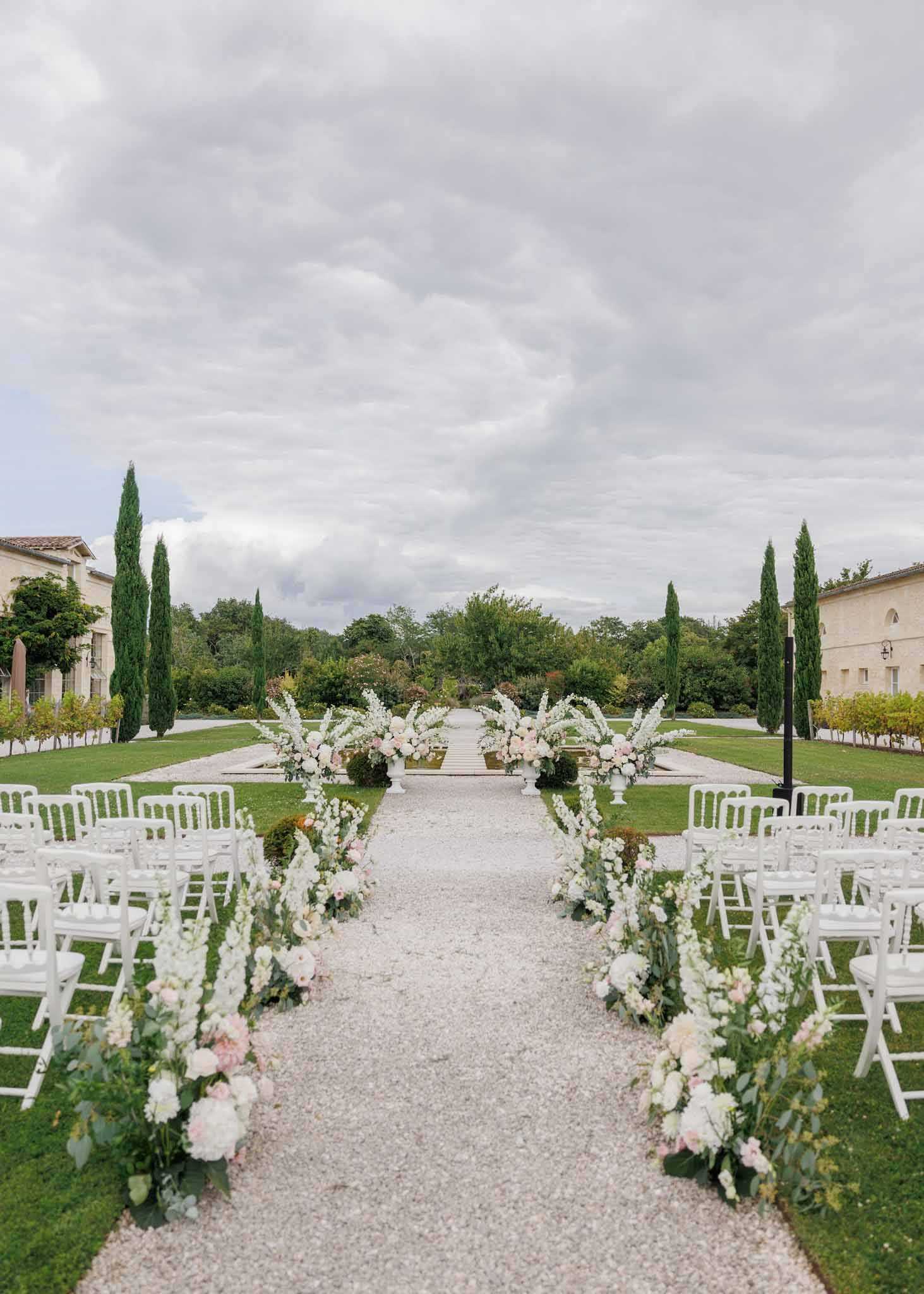 An outdoor wedding ceremony setup at a French estate or chateau, photographed as a wide symmetrical shot looking down the aisle toward a floral altar. The gravel aisle is flanked on both sides by rows of white folding and Chiavari chairs, with lush floral arrangements lining the path at ground level featuring white delphiniums, blush and white peonies, blush roses, and eucalyptus greenery. At the far end, large white urn pedestals hold full, overflowing arrangements in the same white and blush palette. The ceremony space is framed by the stone facades of two venue buildings on either side and tall Italian cypress trees. The overall decor palette is white and soft blush with green foliage accents, giving a classic French garden aesthetic. Potential venue feature image.