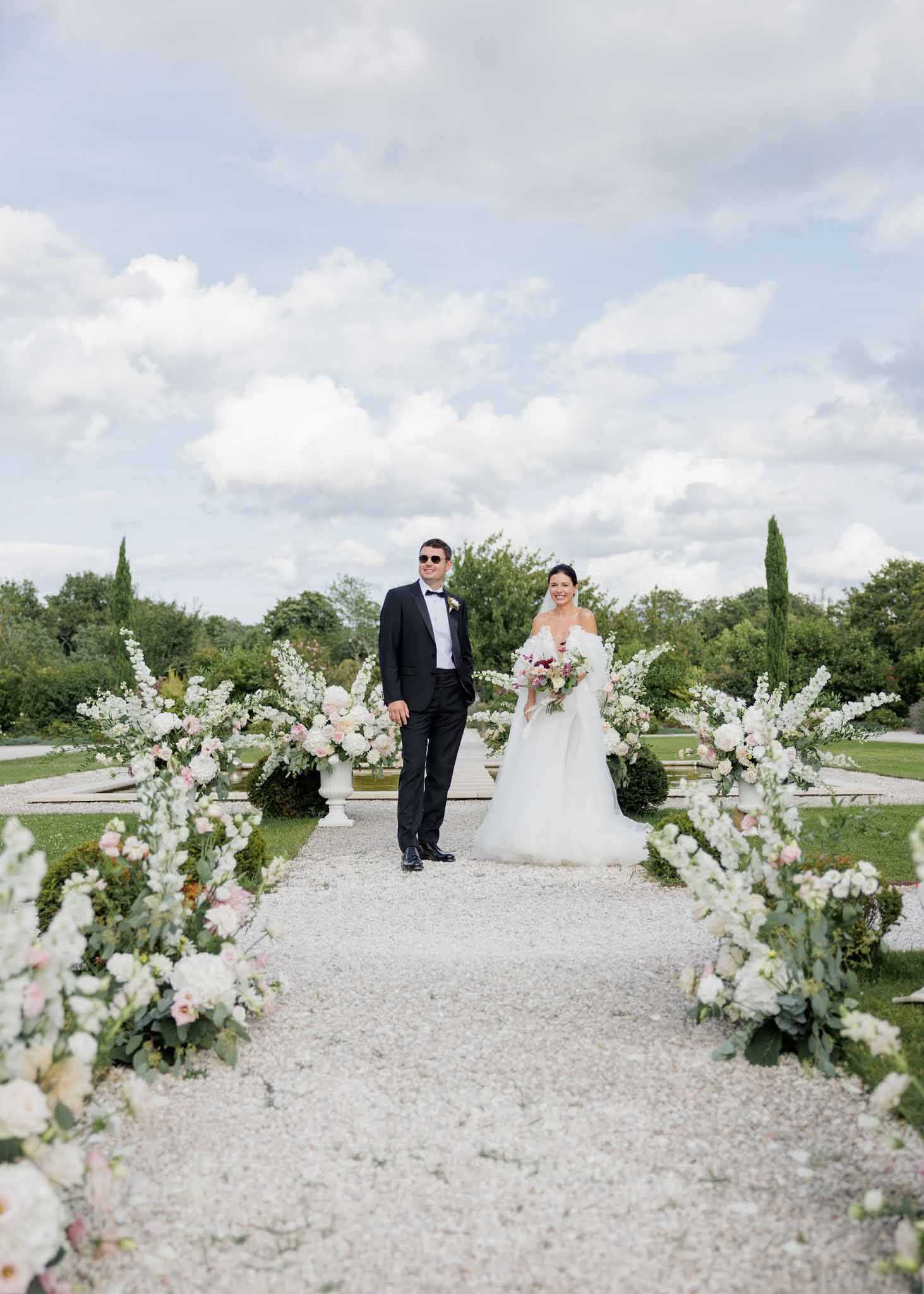 A couple portrait taken outdoors in a formal French garden, with the bride and groom standing together on a gravel pathway flanked by large freestanding floral arrangements. The groom wears a black tuxedo with a bow tie and sunglasses, and the bride wears an off-the-shoulder white ball gown with a tulle skirt, holding a bouquet of white, blush pink, and deep burgundy blooms. The ceremony aisle is lined with lush ground-level floral clusters and tall arrangements in white urns, featuring white hydrangeas, white delphiniums, blush roses, and greenery in a classic white-and-blush palette. The composition is a wide portrait shot centered on the couple with the floral-lined path drawing the eye toward them, set against a formal garden backdrop with trimmed hedges and Italian cypress trees.