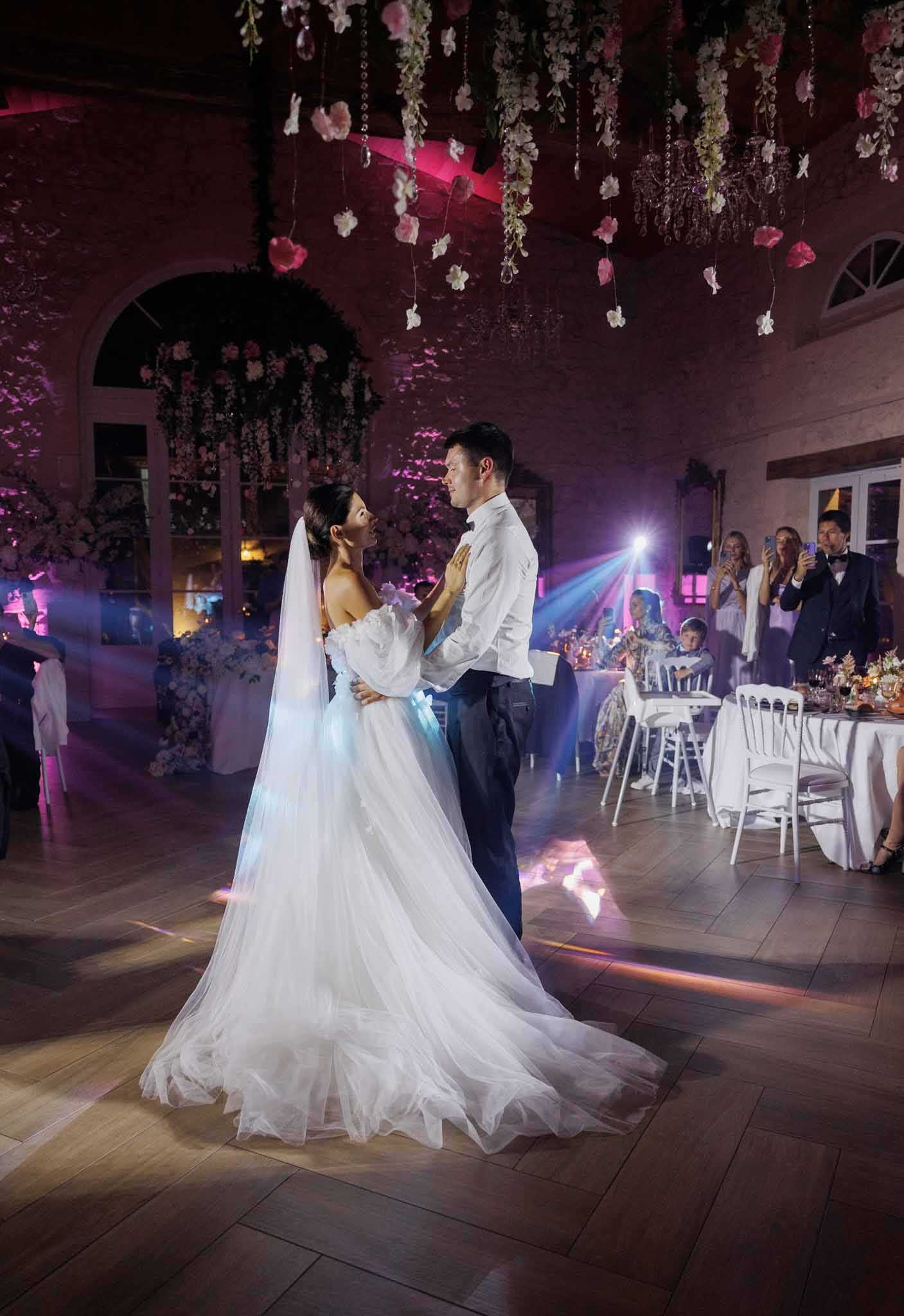 The bride and groom share their first dance in a large indoor reception hall with stone walls, arched windows, and a wood-plank floor. The bride wears an off-the-shoulder white tulle ball gown with long puff sleeves and a cathedral-length veil, while the groom is dressed in a white dress shirt and navy trousers. The ceiling is decorated with hanging installations of blush pink and white florals — including roses and small blooms — suspended on crystal bead strands, and the room is bathed in pink and purple uplighting with dramatic event light beams cutting across the dance floor. To the right, approximately eight to ten guests are seated at white-linen-covered tables with white Chiavari chairs, watching and photographing the couple, with candle and floral centerpieces visible on the tables. A large floral installation of blush pink blooms is visible in the background left corner. The shot is a wide full-length portrait taken from behind and slightly to the side of the couple, capturing the full sweep of the dress train.